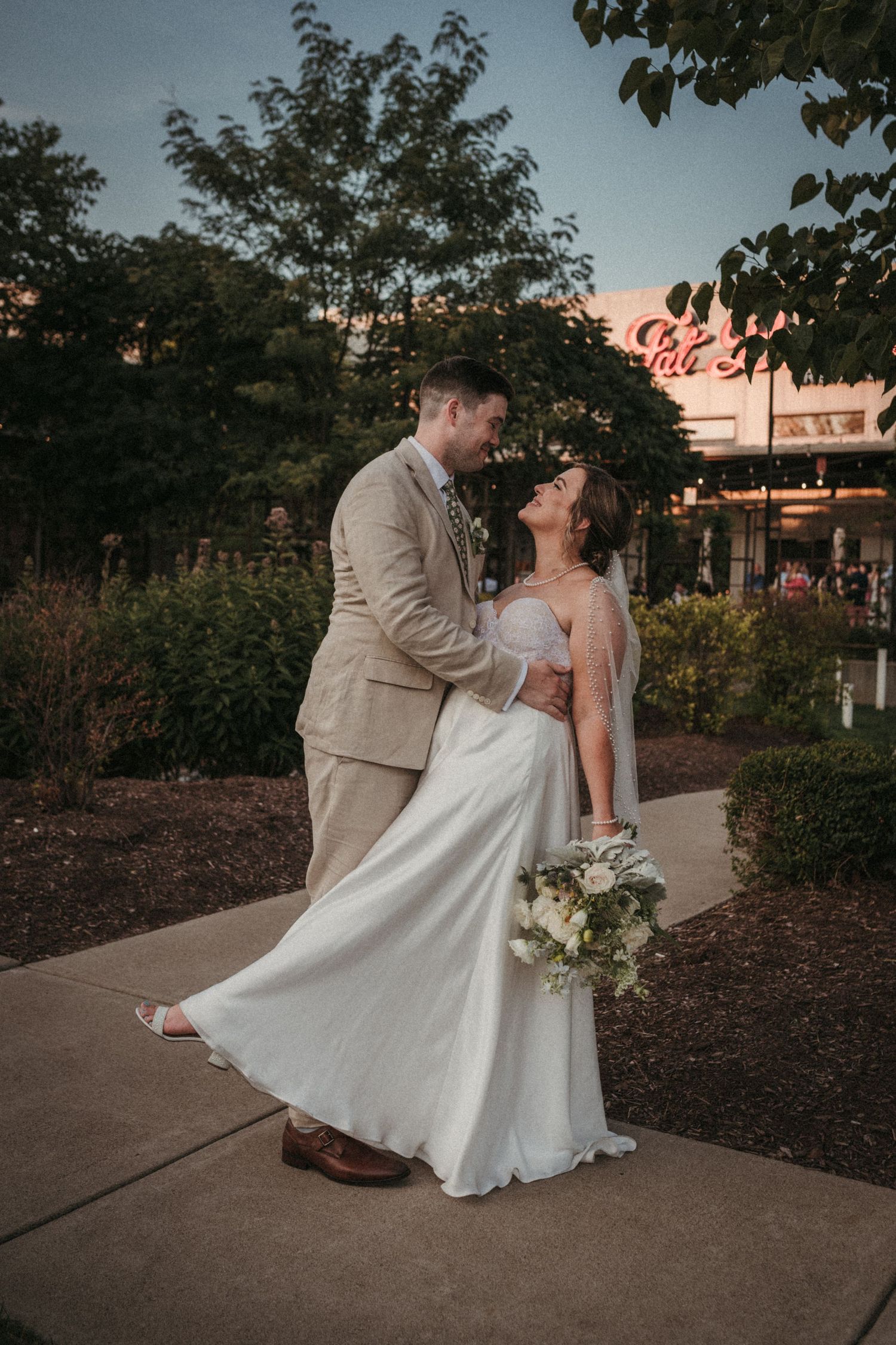 Wedding couple dances together outdoors with bride in white dress and groom in beige suit.