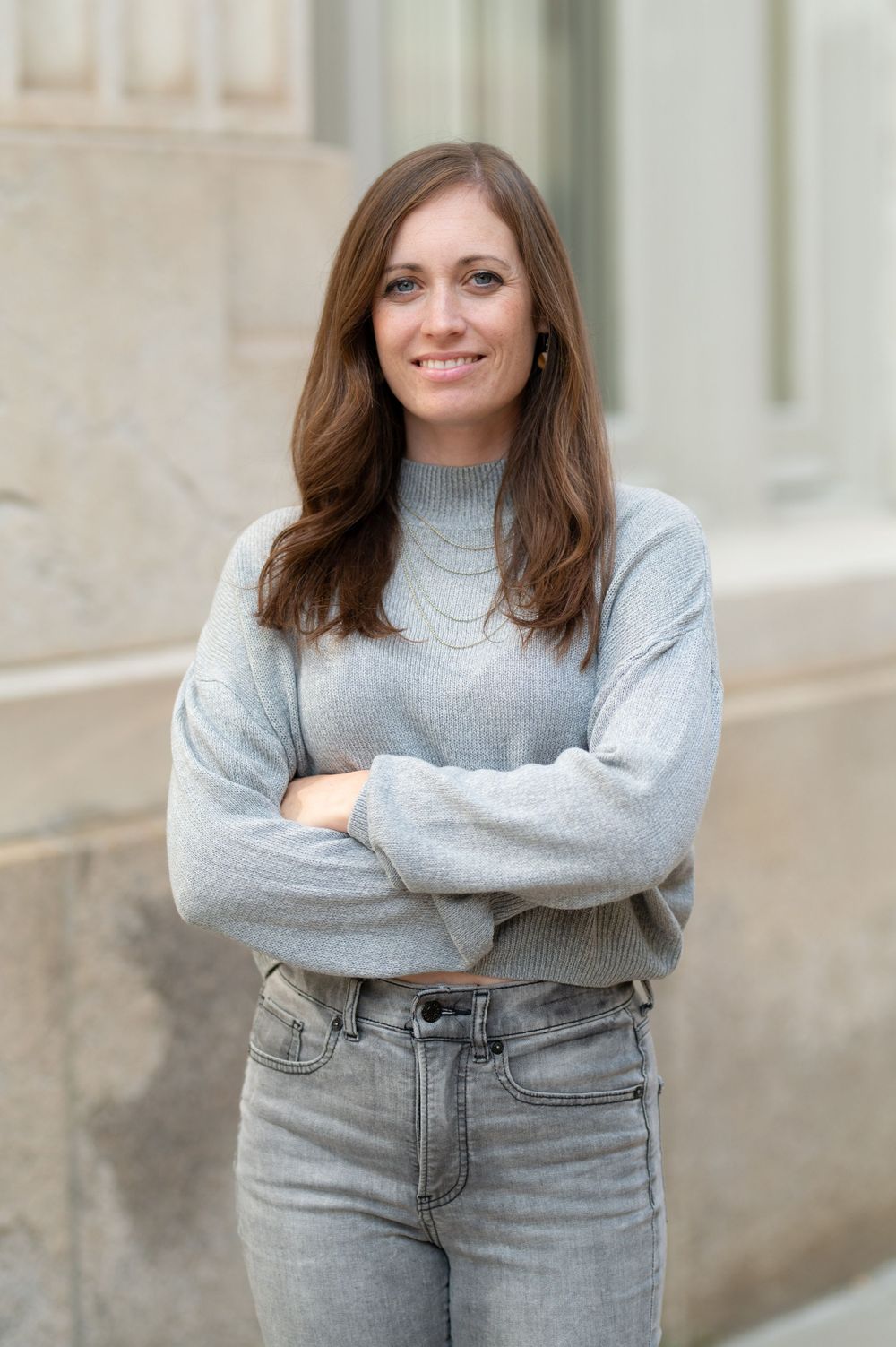 Casual pose with arms crossed wearing gray sweater against stone building.