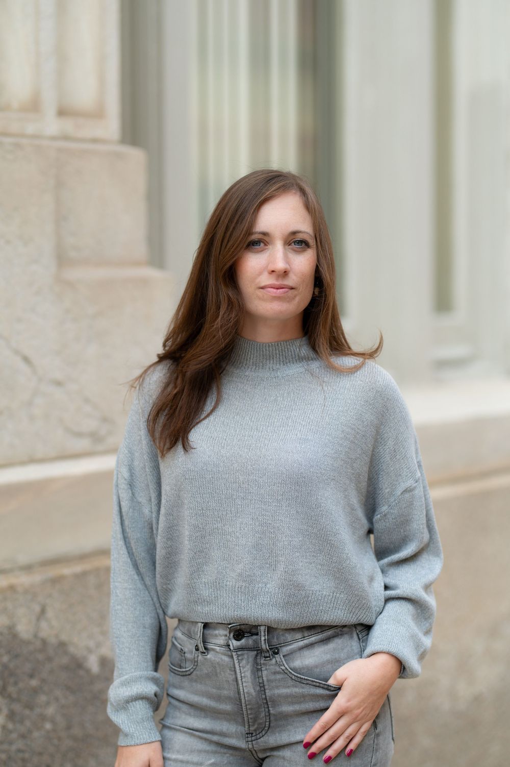 Person wearing a light gray sweater and jeans posing against a stone building exterior.