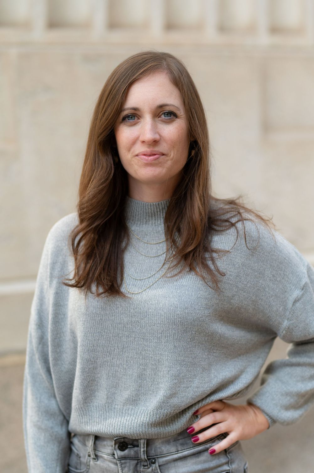 A portrait of someone wearing a light gray sweater against a beige architectural backdrop.