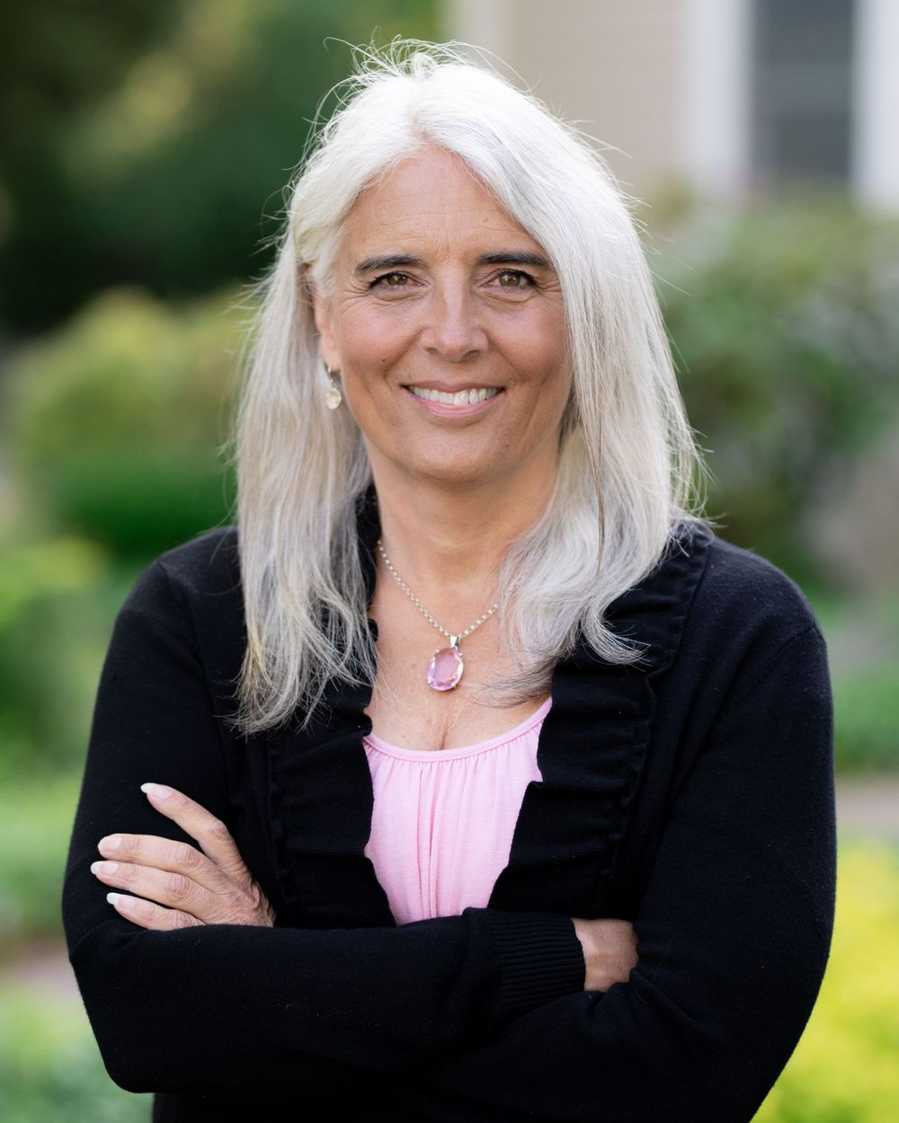 Smiling portrait in black cardigan and pink top against garden backdrop.
