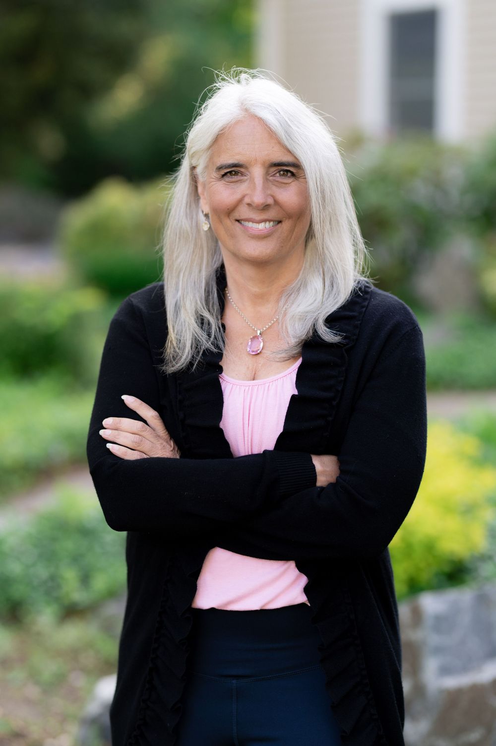Confident pose in black cardigan and pink top with garden background.