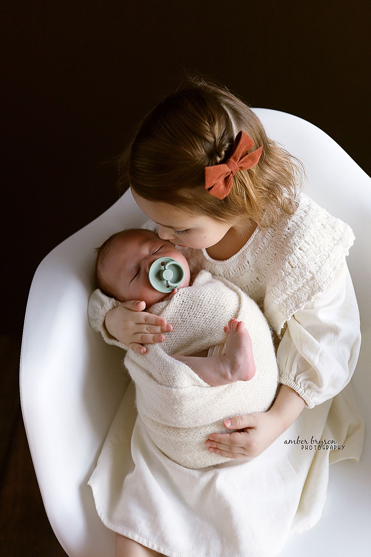 A tender moment between parent and infant wrapped in white, sharing an intimate embrace against a dark background.