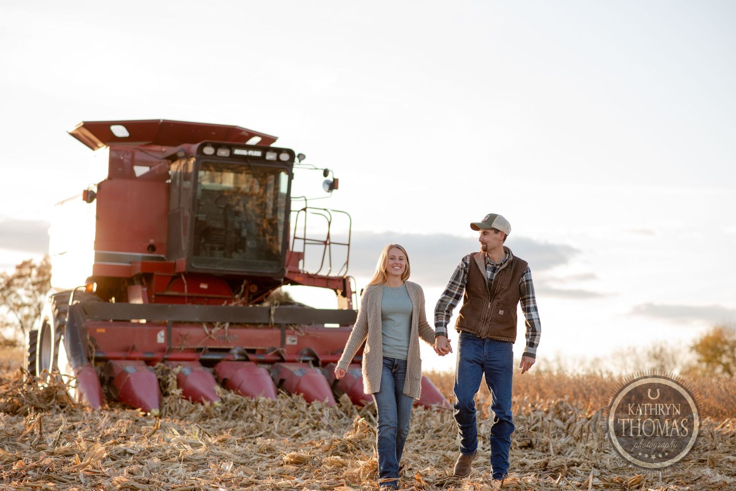 Couple walking together near a large red combine harvester in a farm field at sunset.