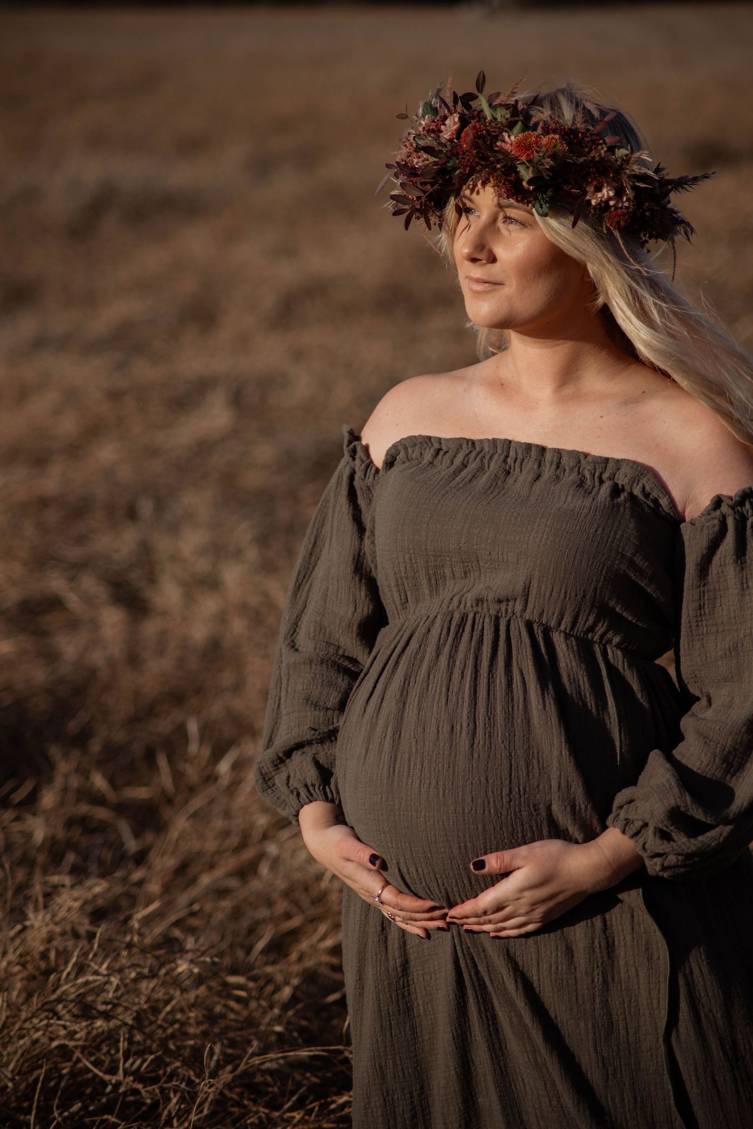Pregnant woman in olive off-shoulder dress and floral crown poses in golden field at sunset.