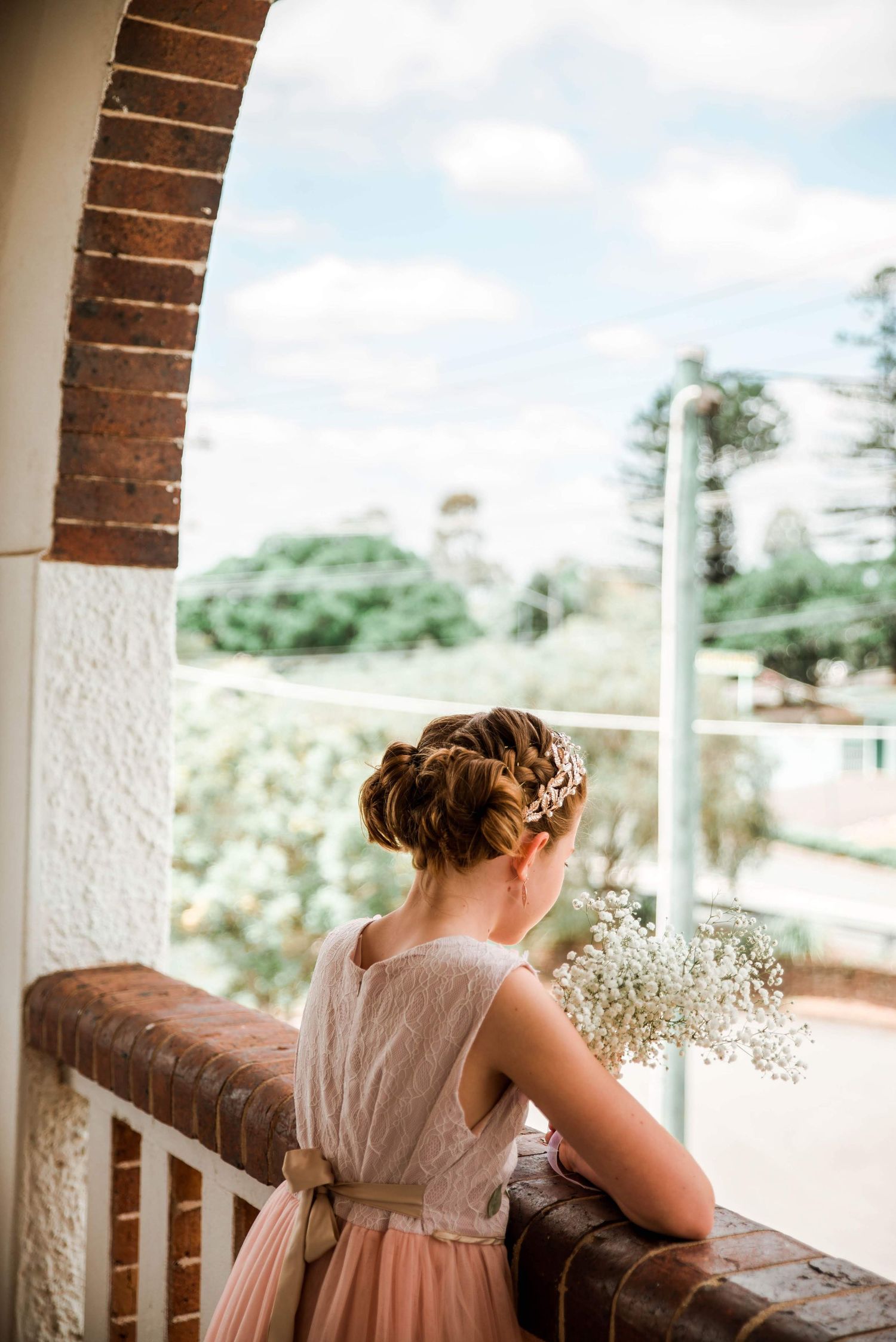 Person in vintage-style dress standing on balcony holding baby's breath flowers looking at scenic view.