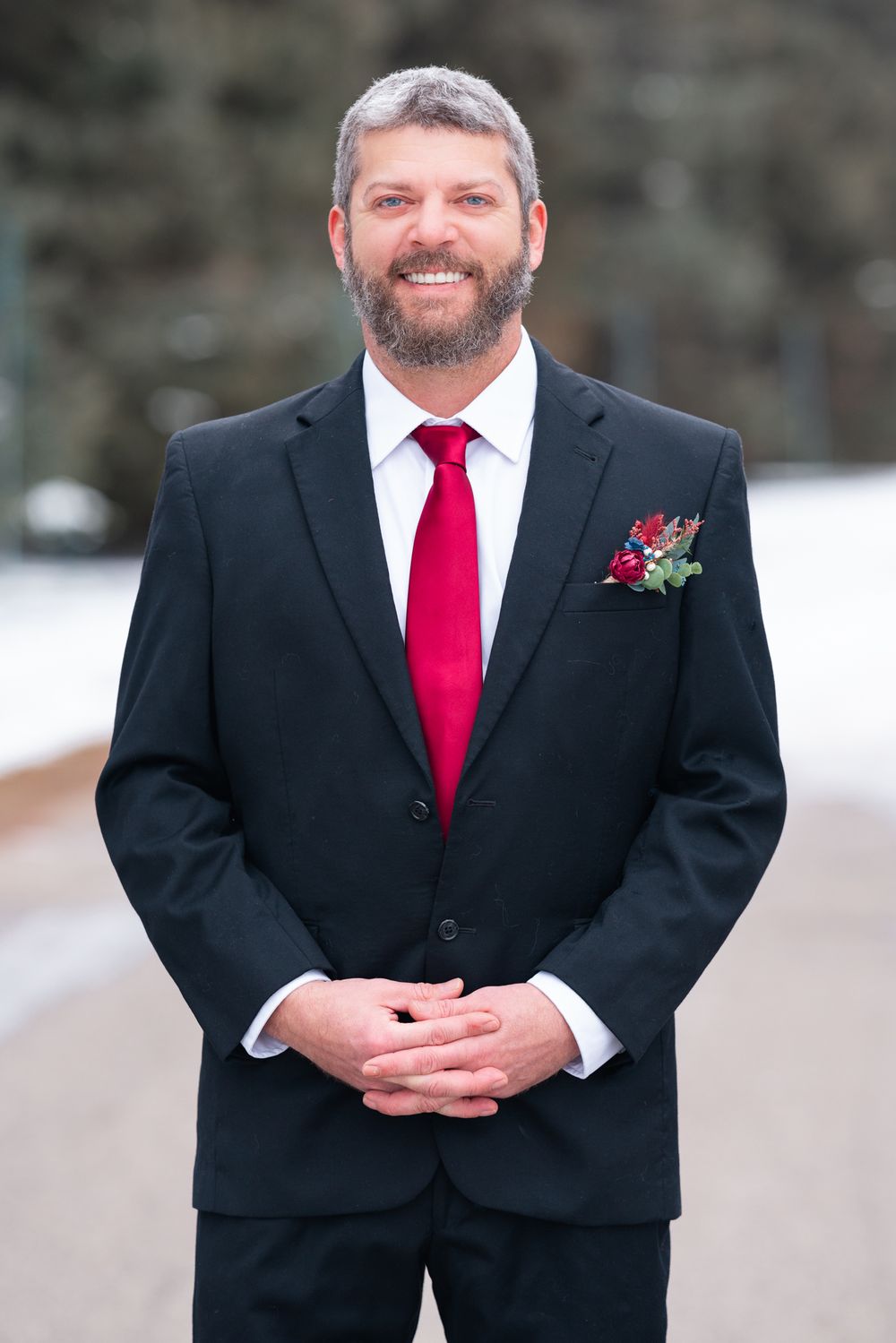 Groom in black suit with burgundy tie and boutonniere smiles at winter wedding.