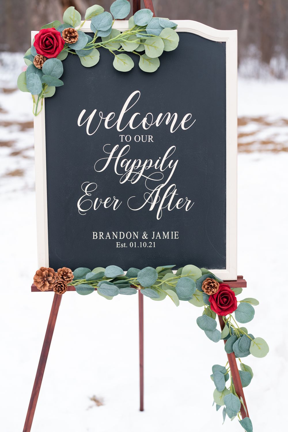 Elegant wedding welcome sign with eucalyptus and red rose decorations on a black chalkboard.