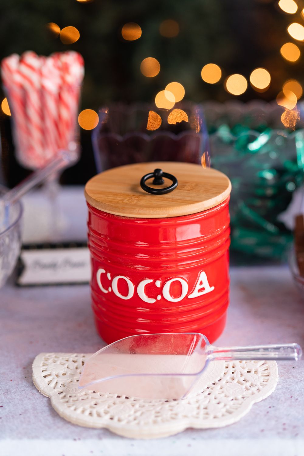 Red ceramic cocoa container with candy canes and holiday bokeh lights in background.