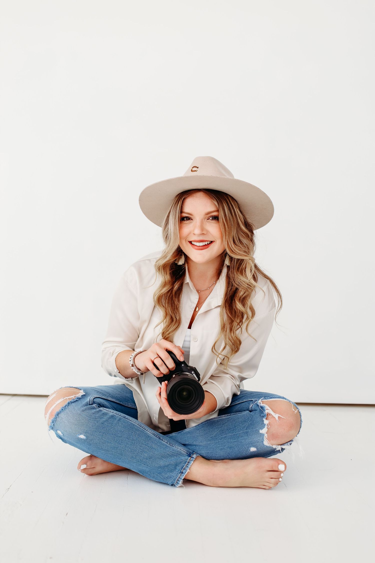Smiling photographer in casual attire sitting cross-legged with camera against white background.
