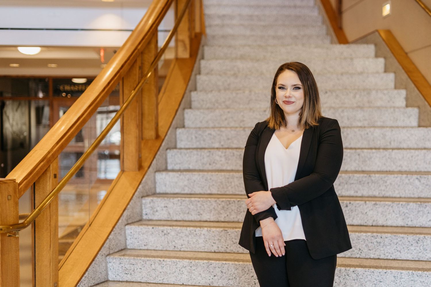 Professional in business attire stands confidently on marble staircase with wooden railing in modern office setting.
