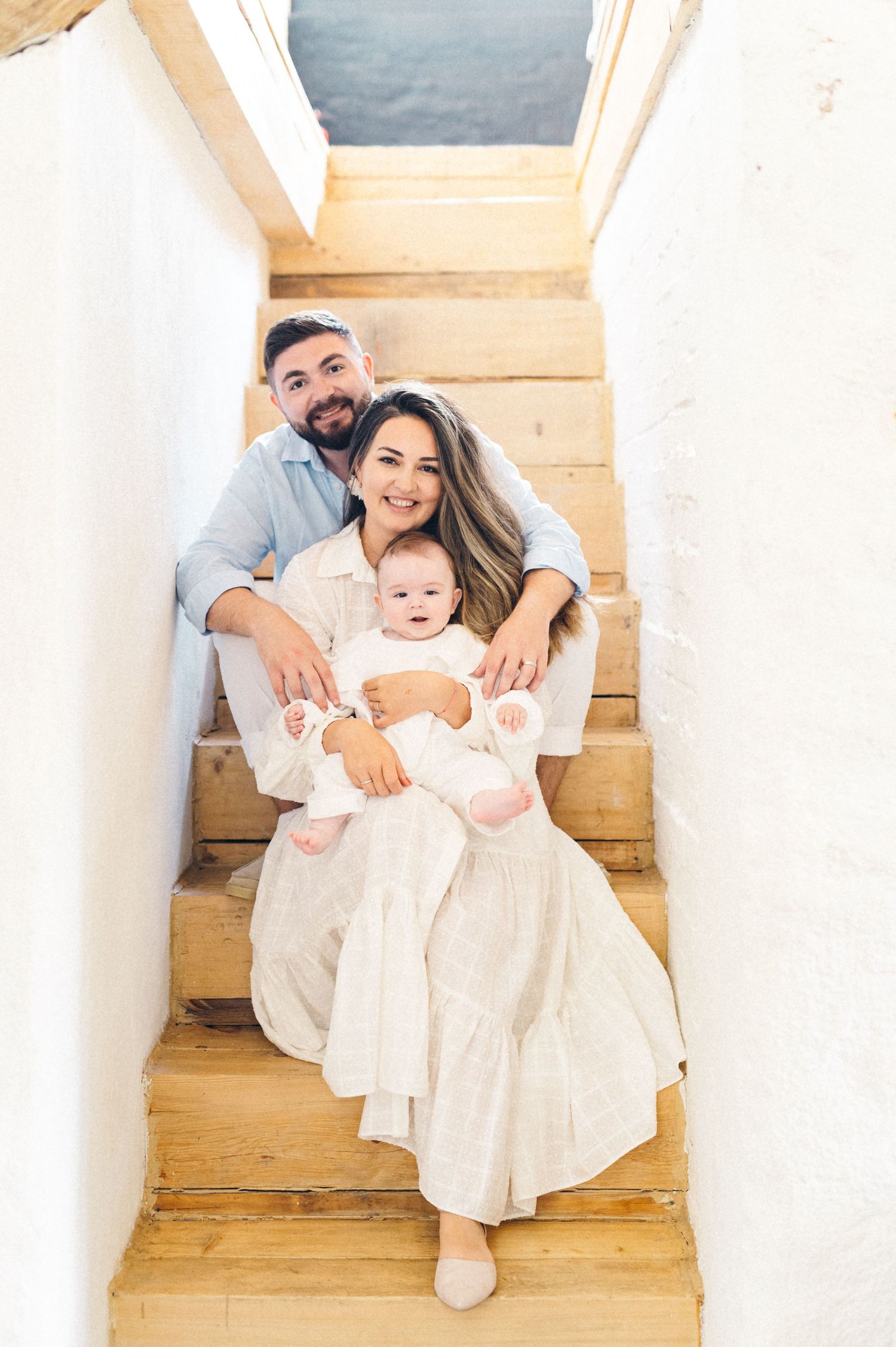 Family in white outfits sits together on wooden stairs in an intimate portrait setting.