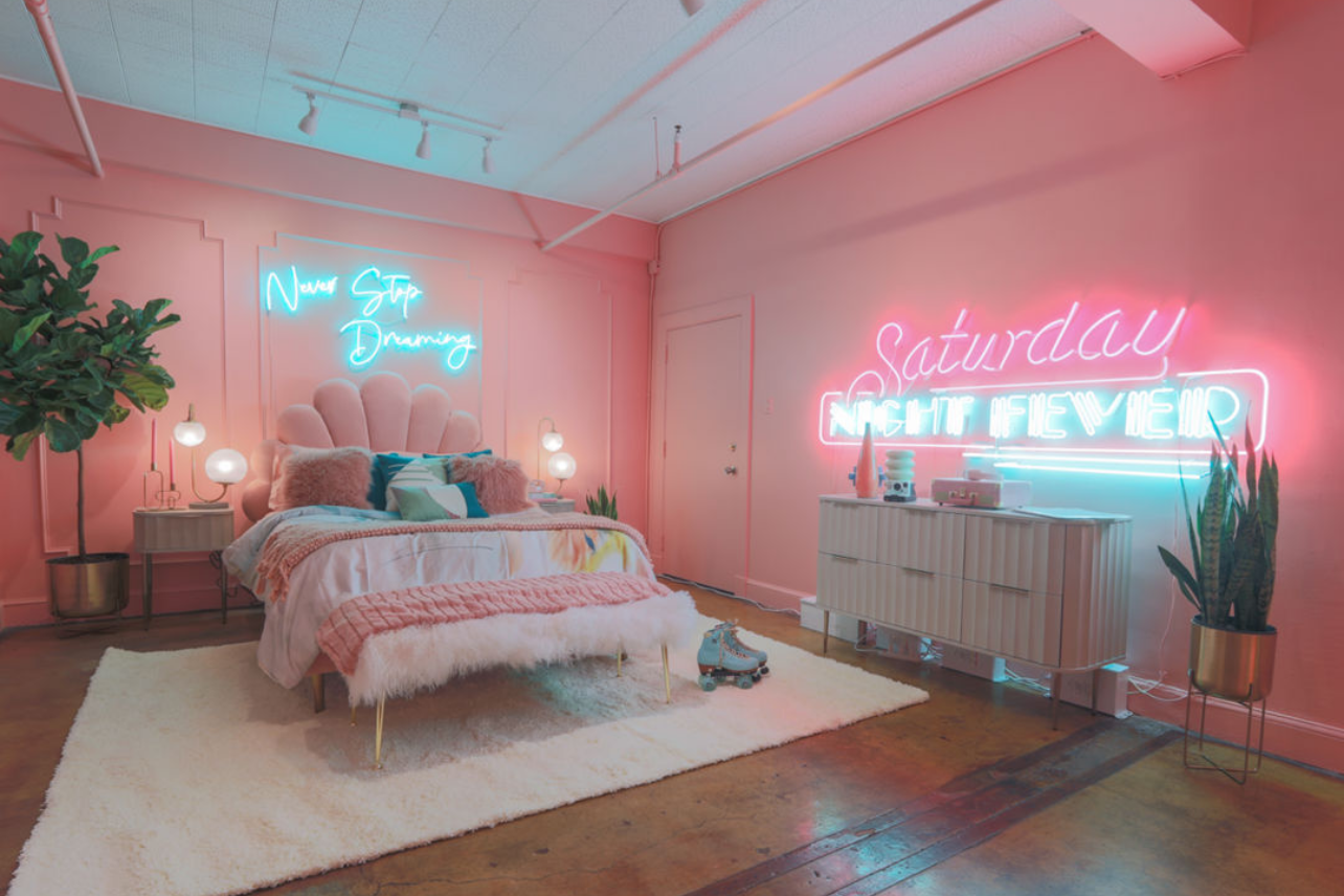 Pink bedroom with neon signs, white bedding, and plants creating a dreamy aesthetic.