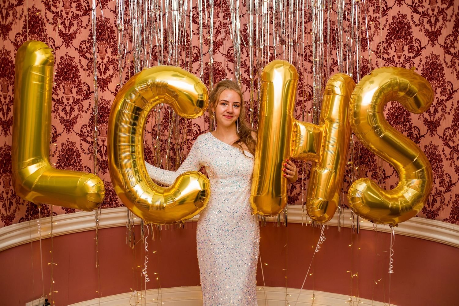 Party scene with gold balloon letters and sparkly decorations against a red damask wallpaper.