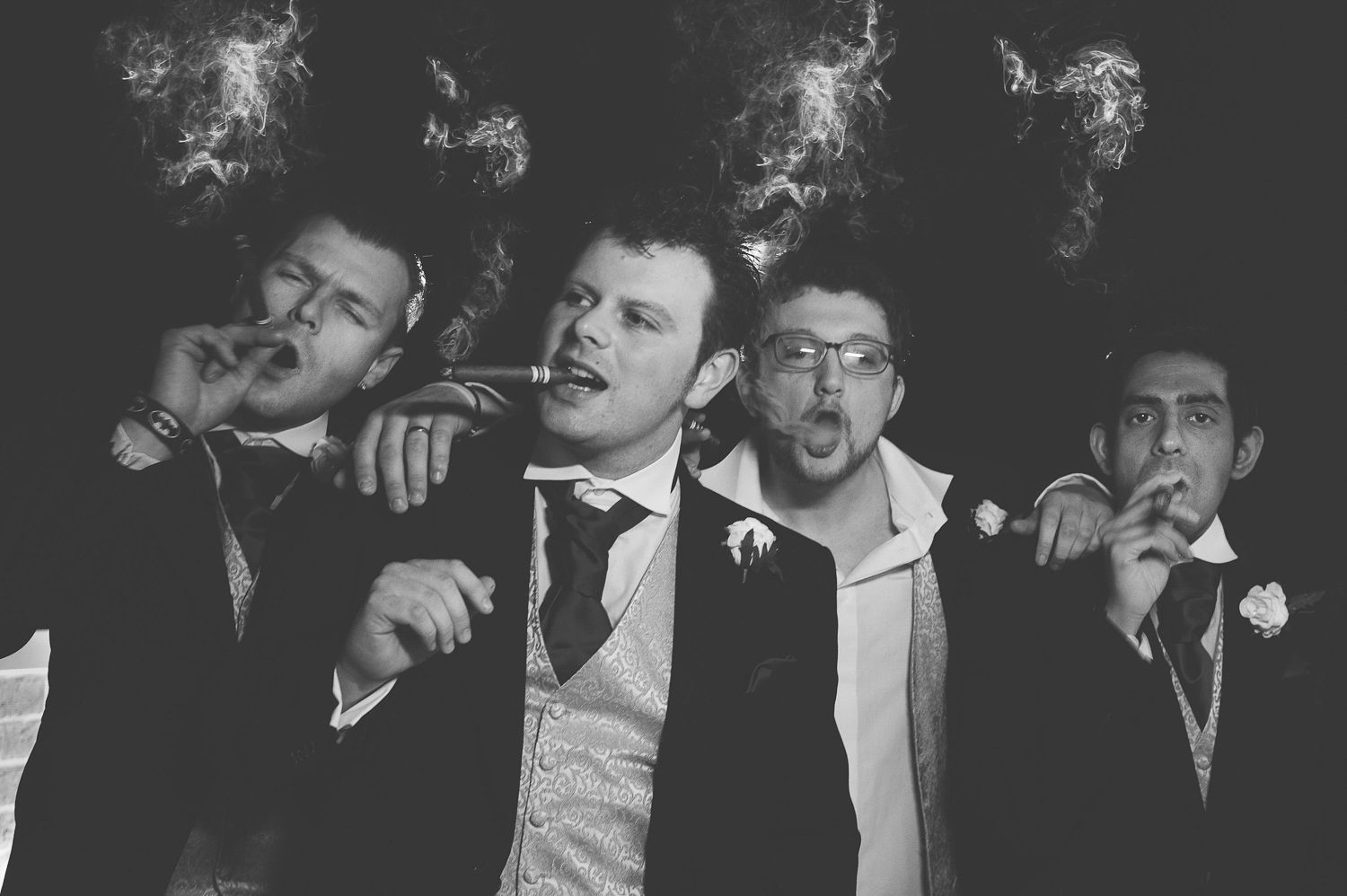 Groomsmen smoke cigars and pose playfully in black and white photography.