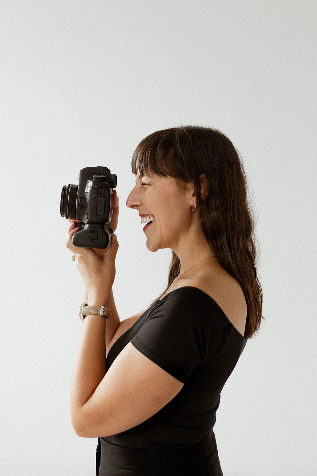 A photographer in black attire smiles while looking at their camera against a white backdrop.