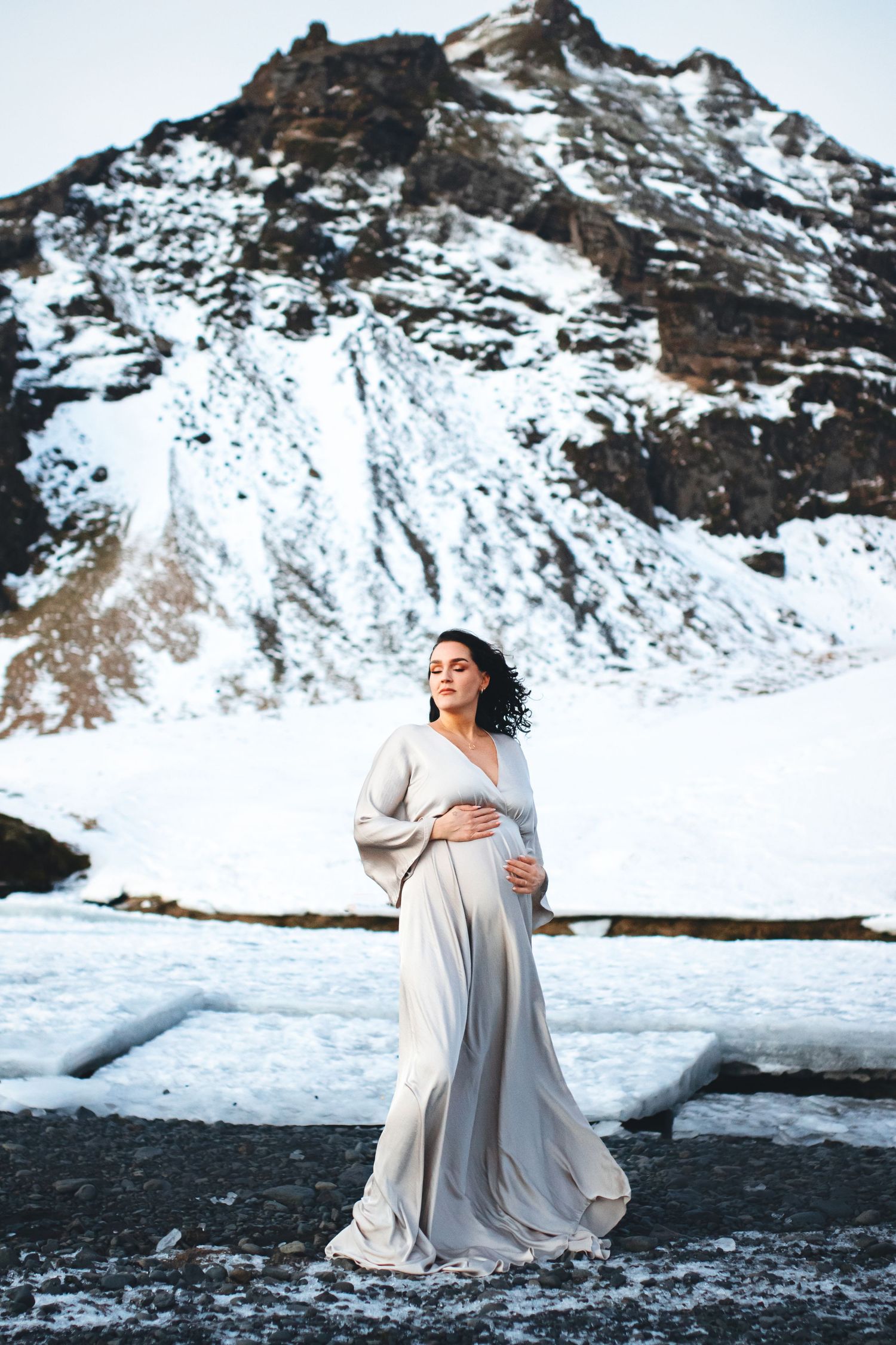 A maternity photo shoot features a woman in flowing dress against snowy mountain backdrop.