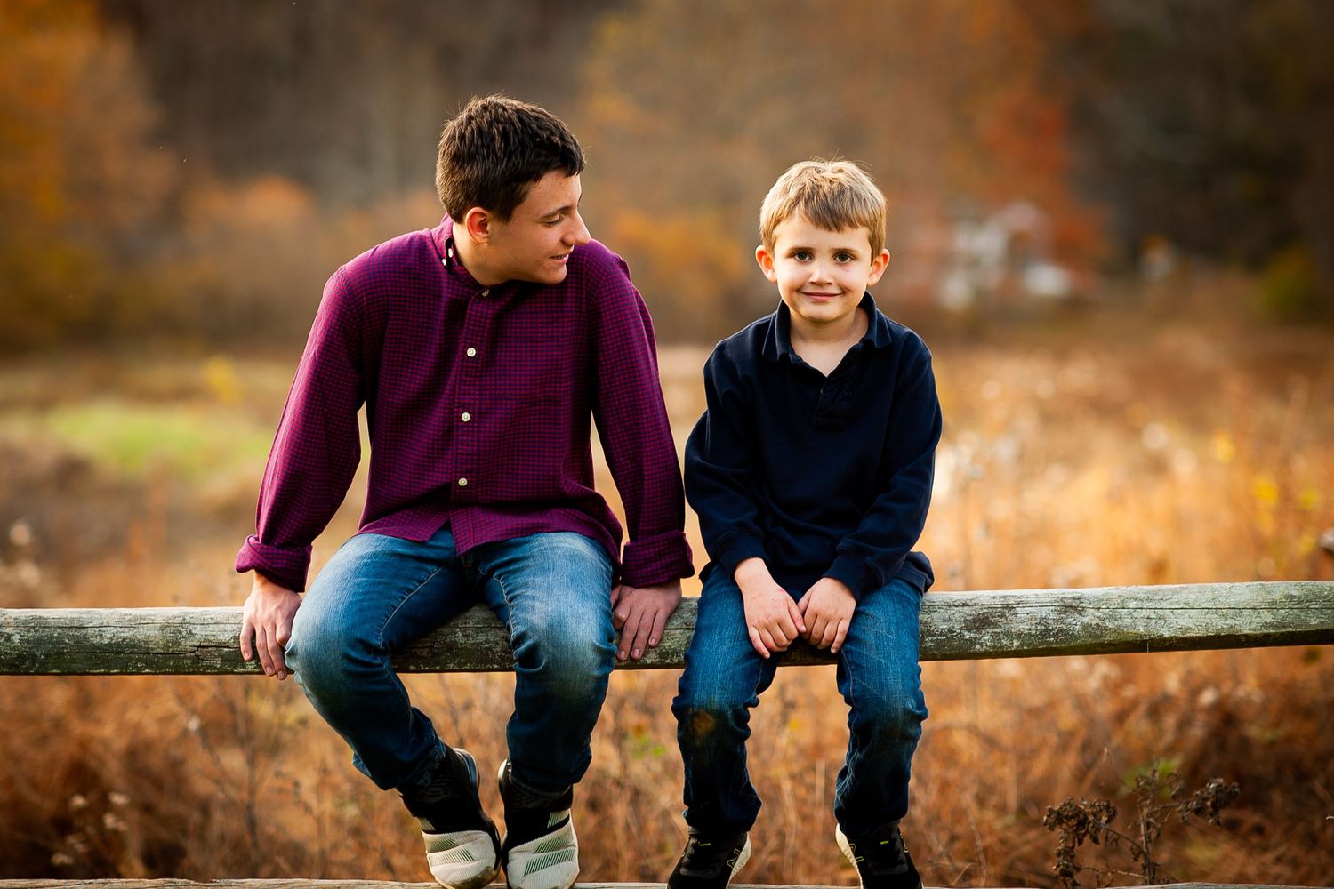 Two boys, seated on a fence with fall backdrop. One looking at his brother, the other one at camera.