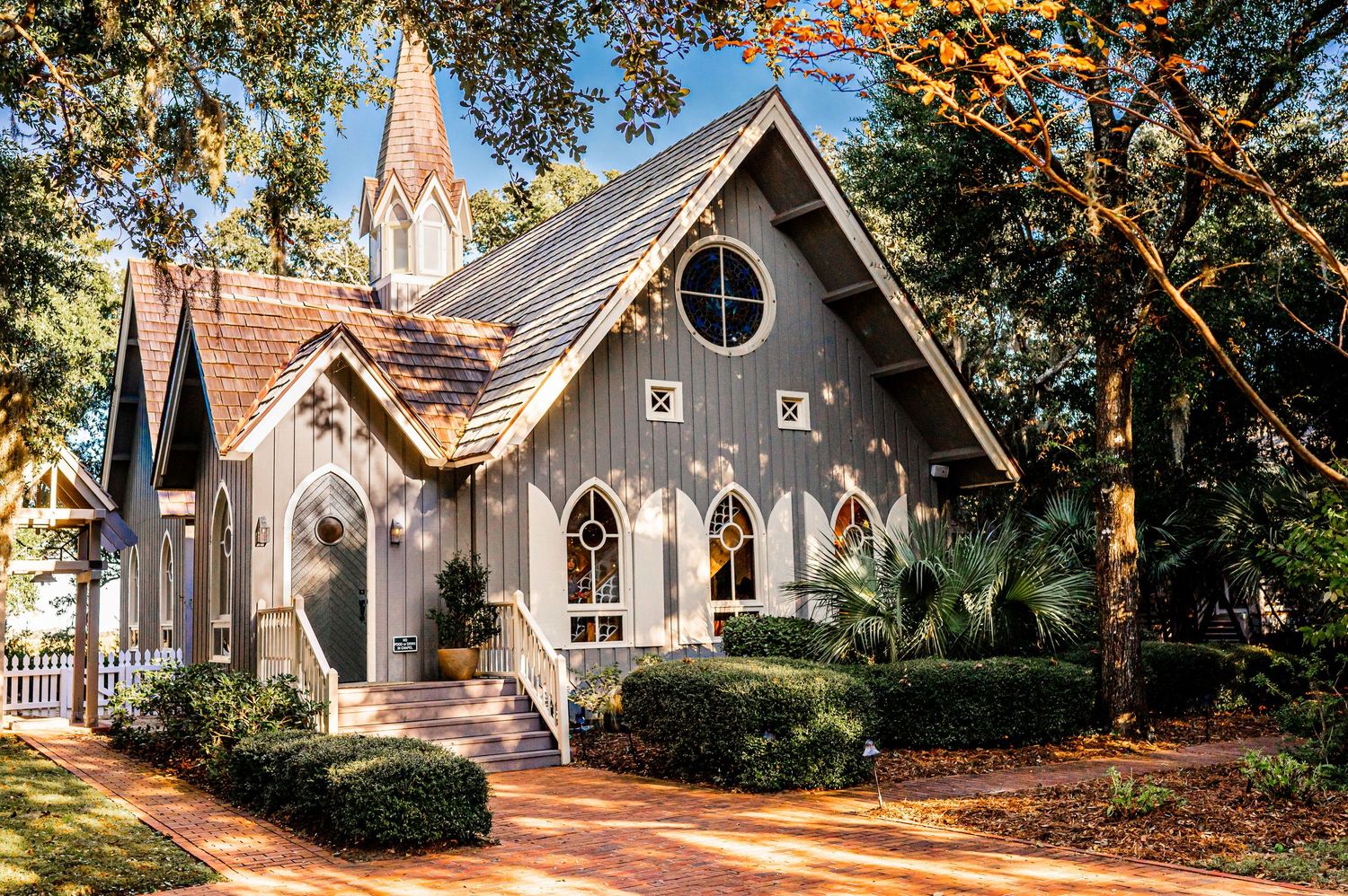 A charming Gothic Revival chapel with gray exterior and pointed arches nestled among trees on a sunny day.