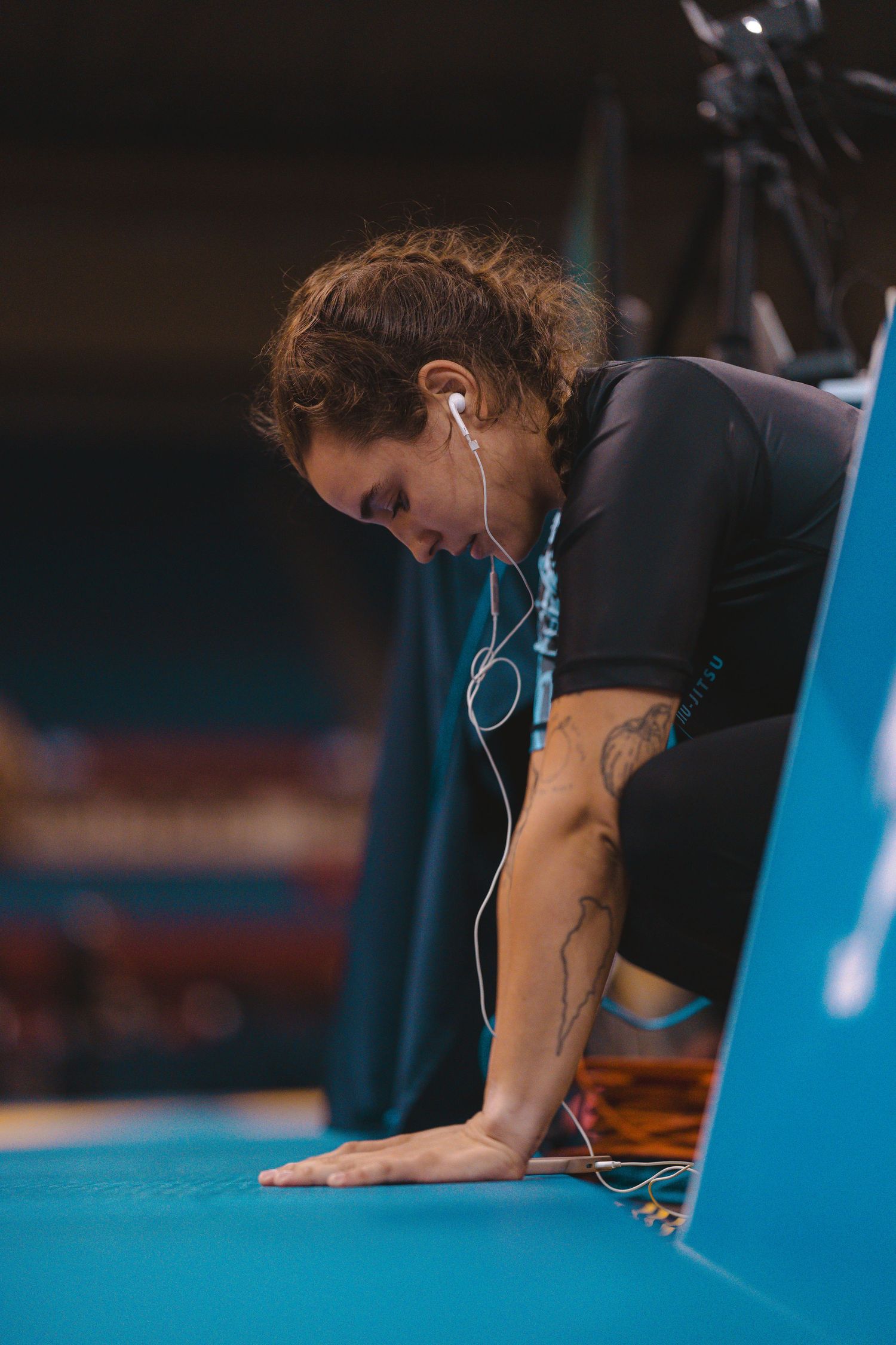 Athlete in black uniform leans forward with earbuds on blue competition surface.
