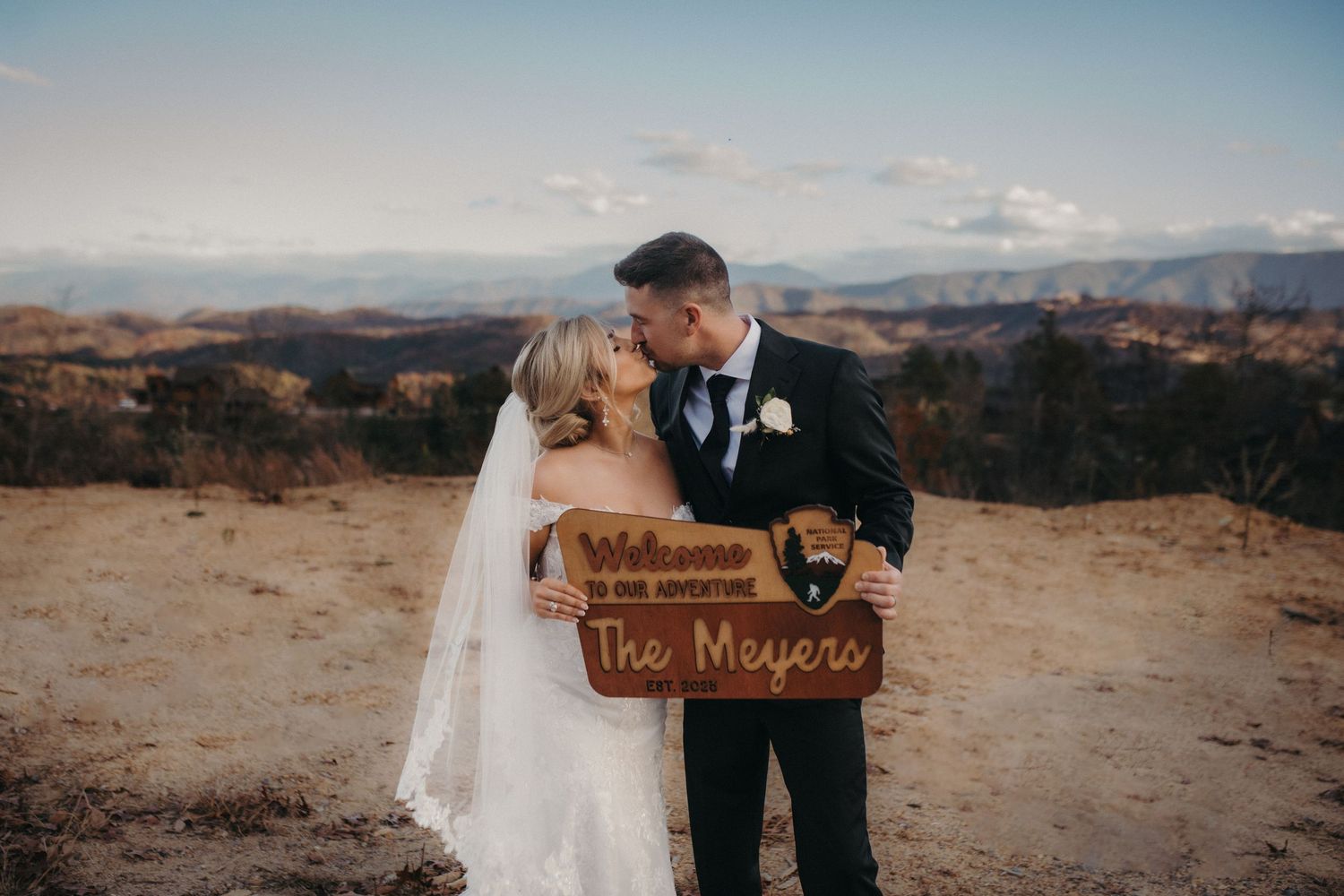 A couple holds a rustic wooden wedding sign against a desert mountain backdrop.
