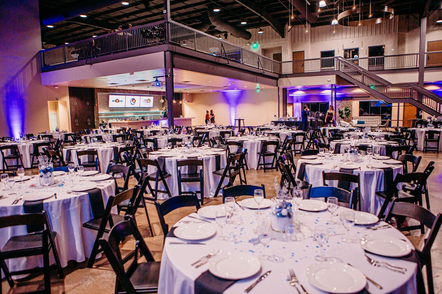 Elegant banquet hall setup with round tables and black chairs under purple ambient lighting.
