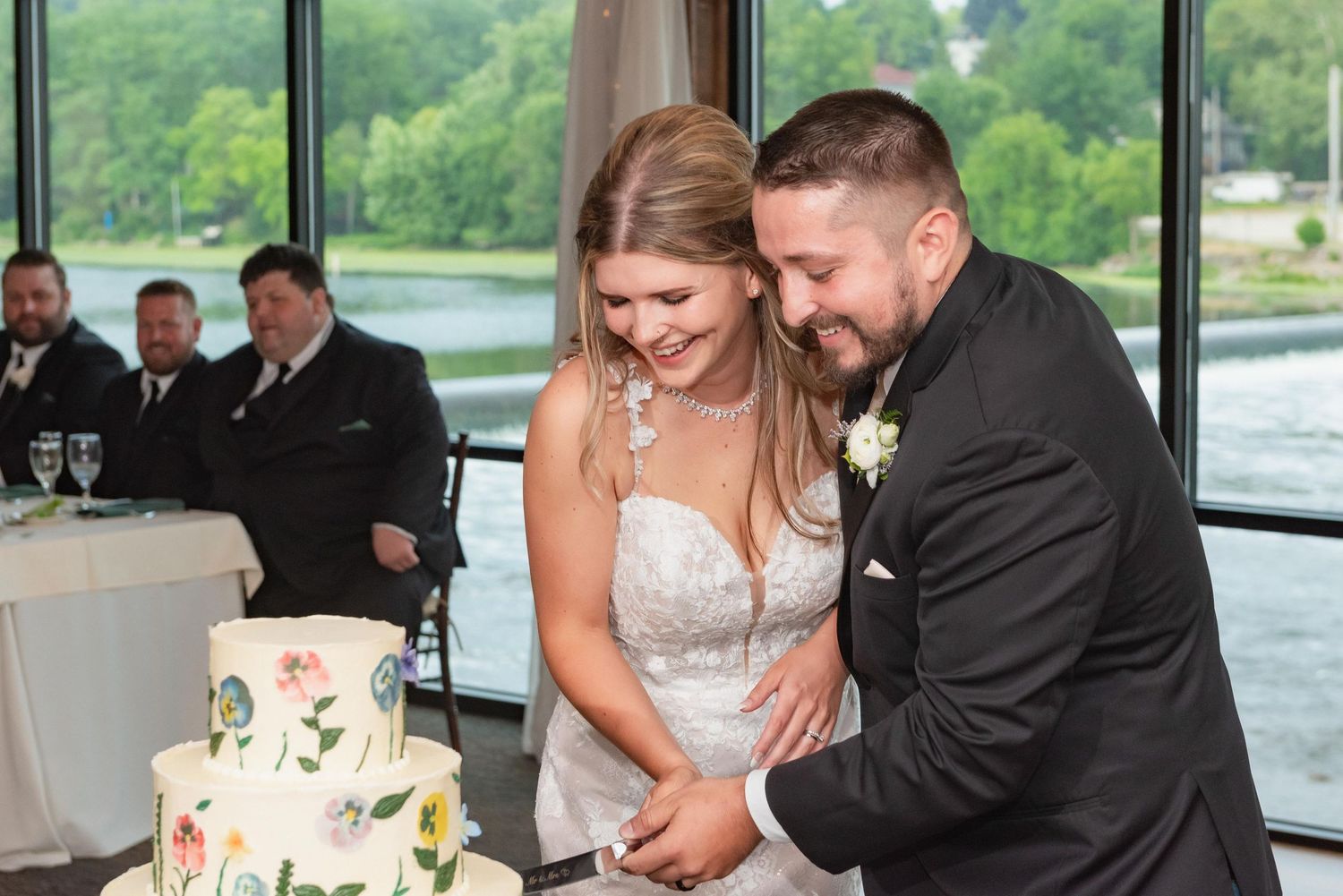 Wedding couple cutting their cake by large windows overlooking water view.