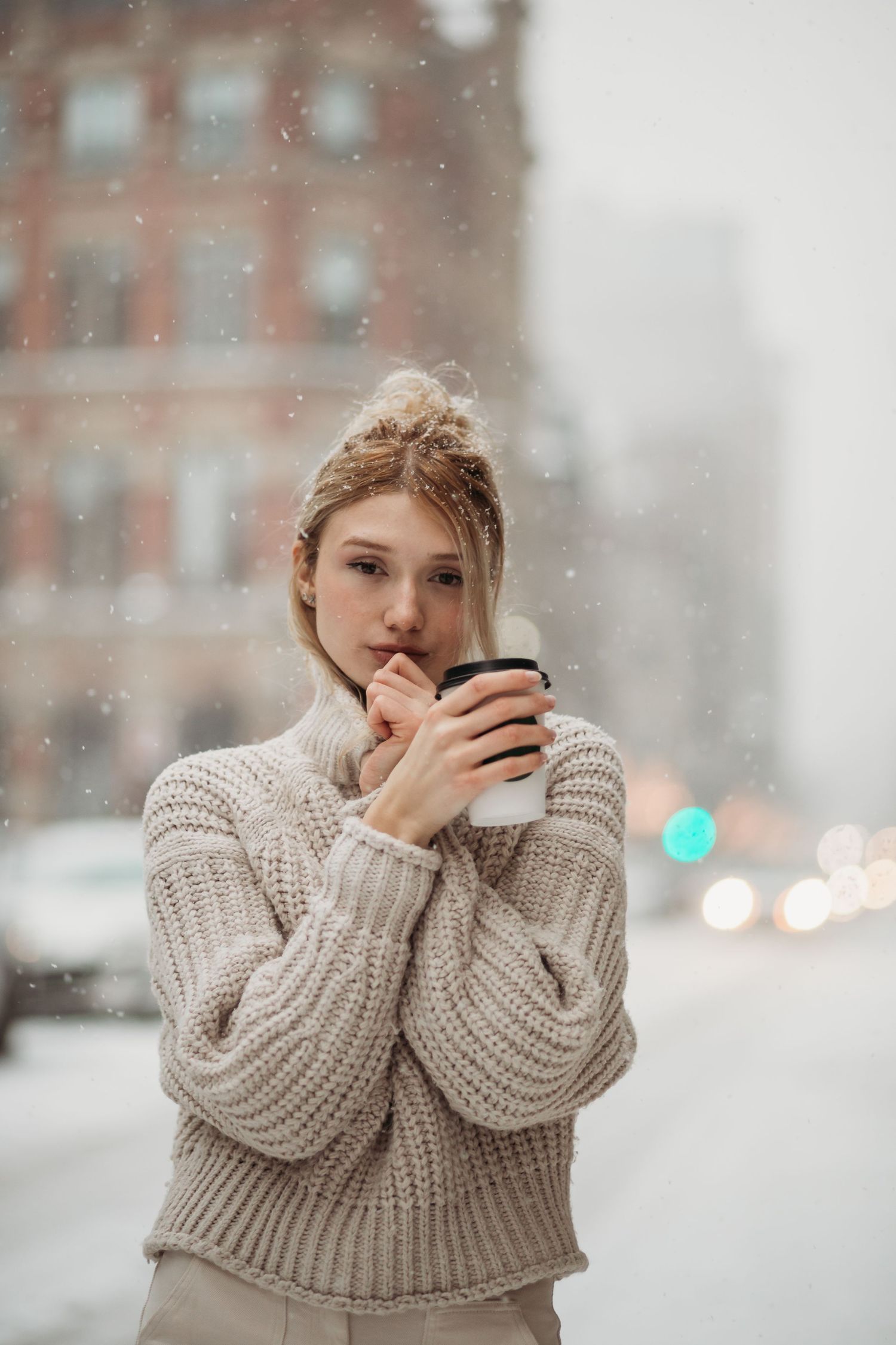 Person in chunky knit sweater holds warm drink while standing in snowy weather with urban backdrop.