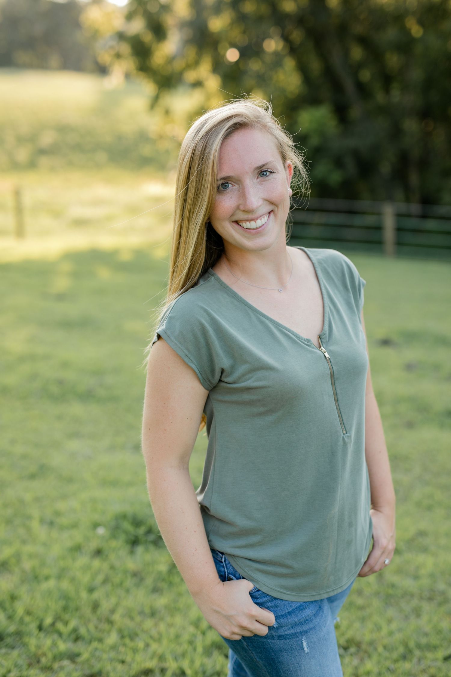 Woman in sage green top smiles in sunny outdoor setting with natural bokeh background.