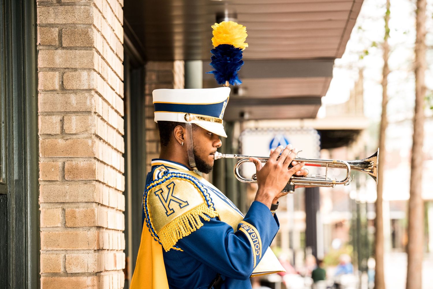 Marching band member plays trumpet while wearing blue and gold uniform.