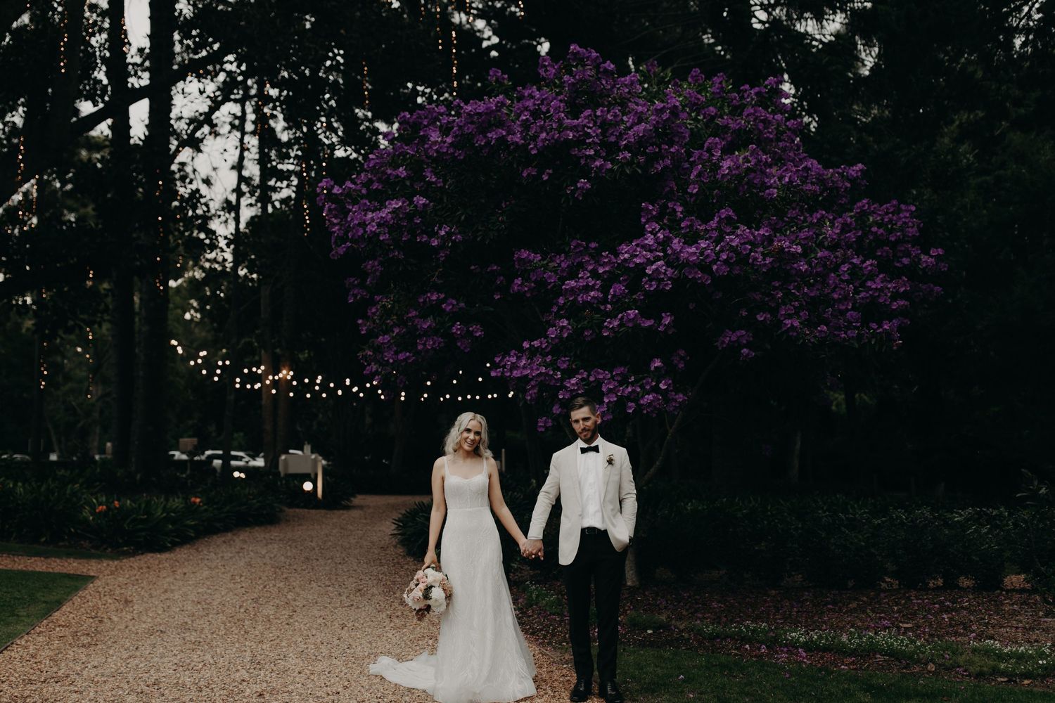Wedding couple walking through garden path with purple flowering trees.