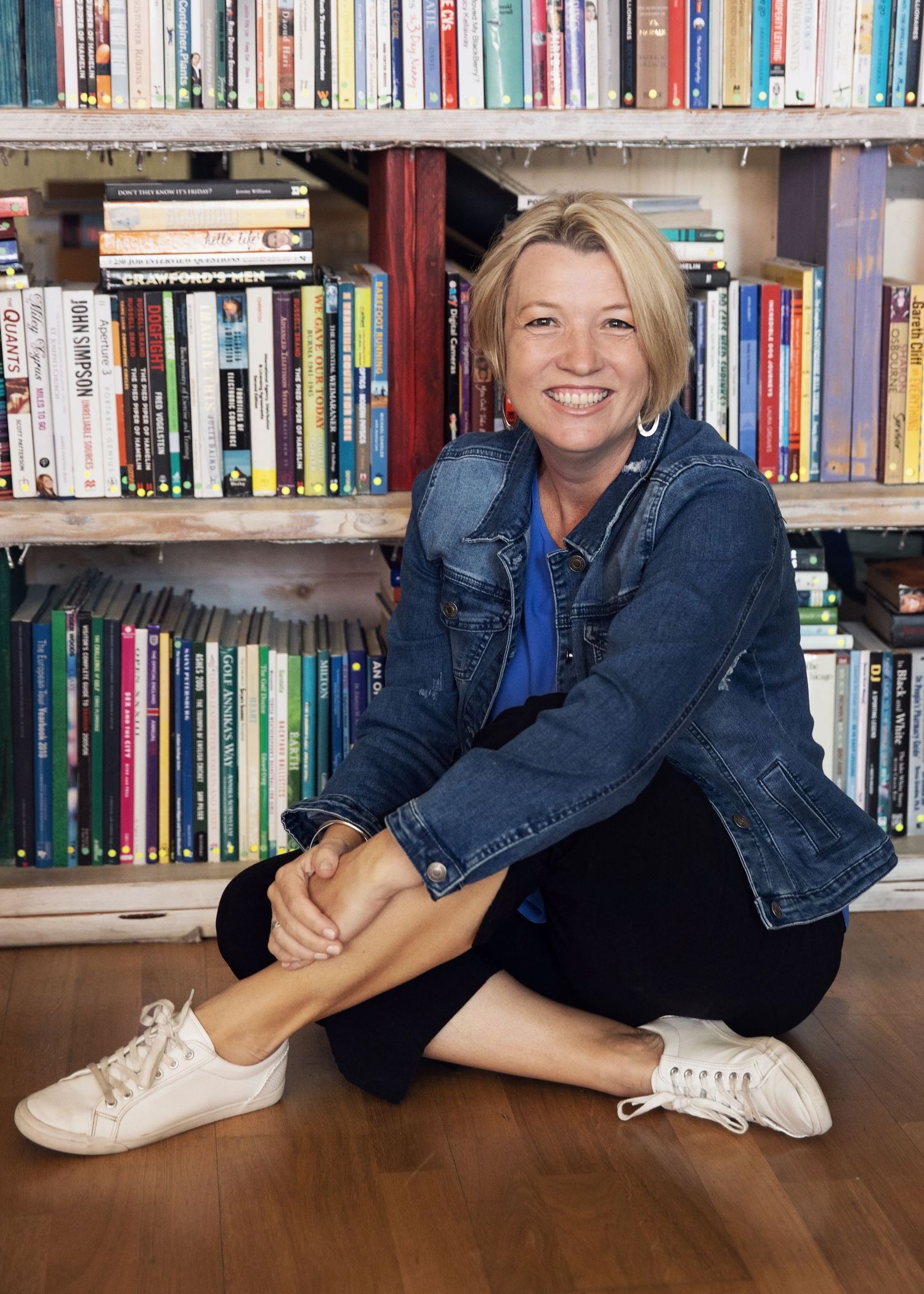 Reader sits cross-legged on wooden floor by bookshelf wearing denim jacket and white sneakers with books lined up behind.