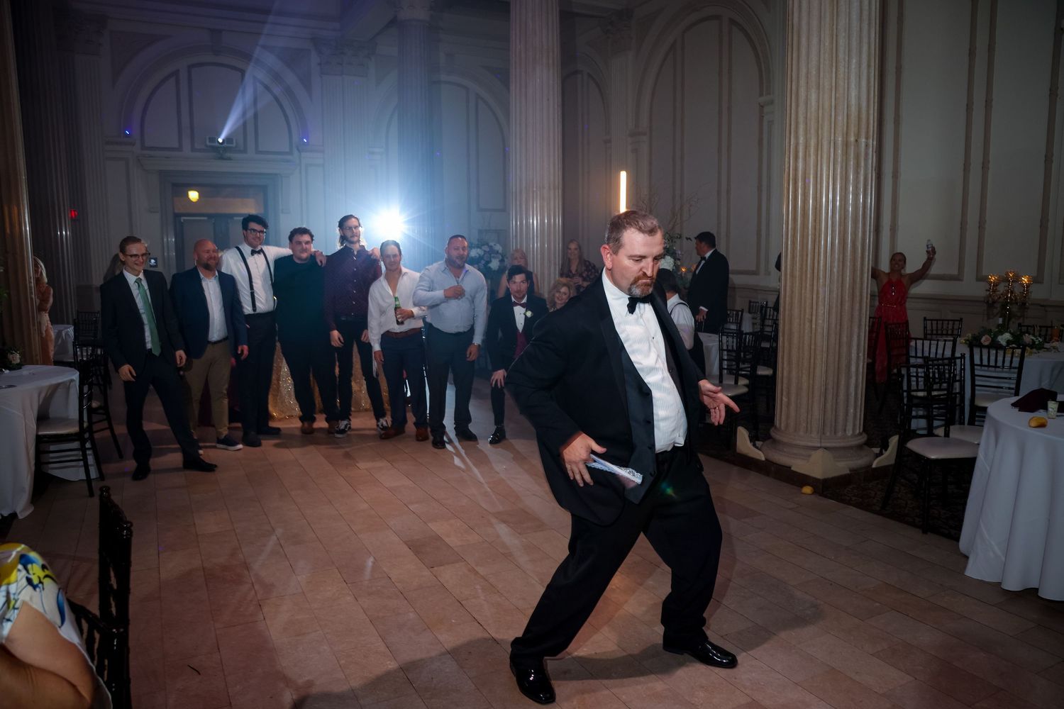 A wedding guest dances enthusiastically in a tuxedo at an elegant reception venue.