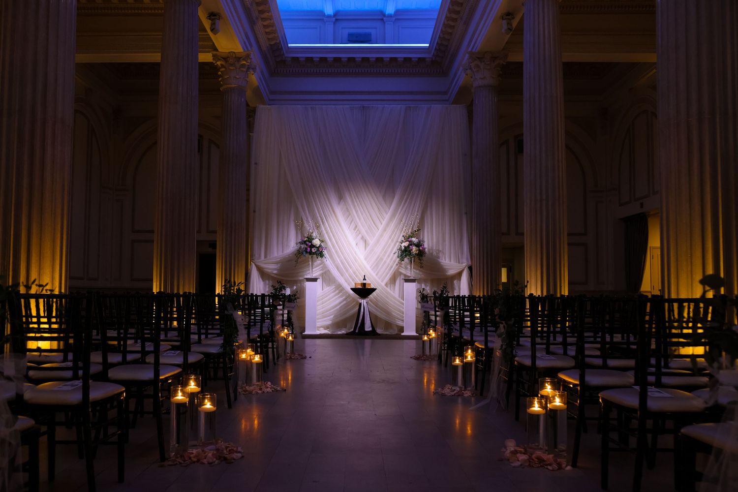 Elegant ceremony space with candlelit aisle and dramatic uplighting on classical columns.