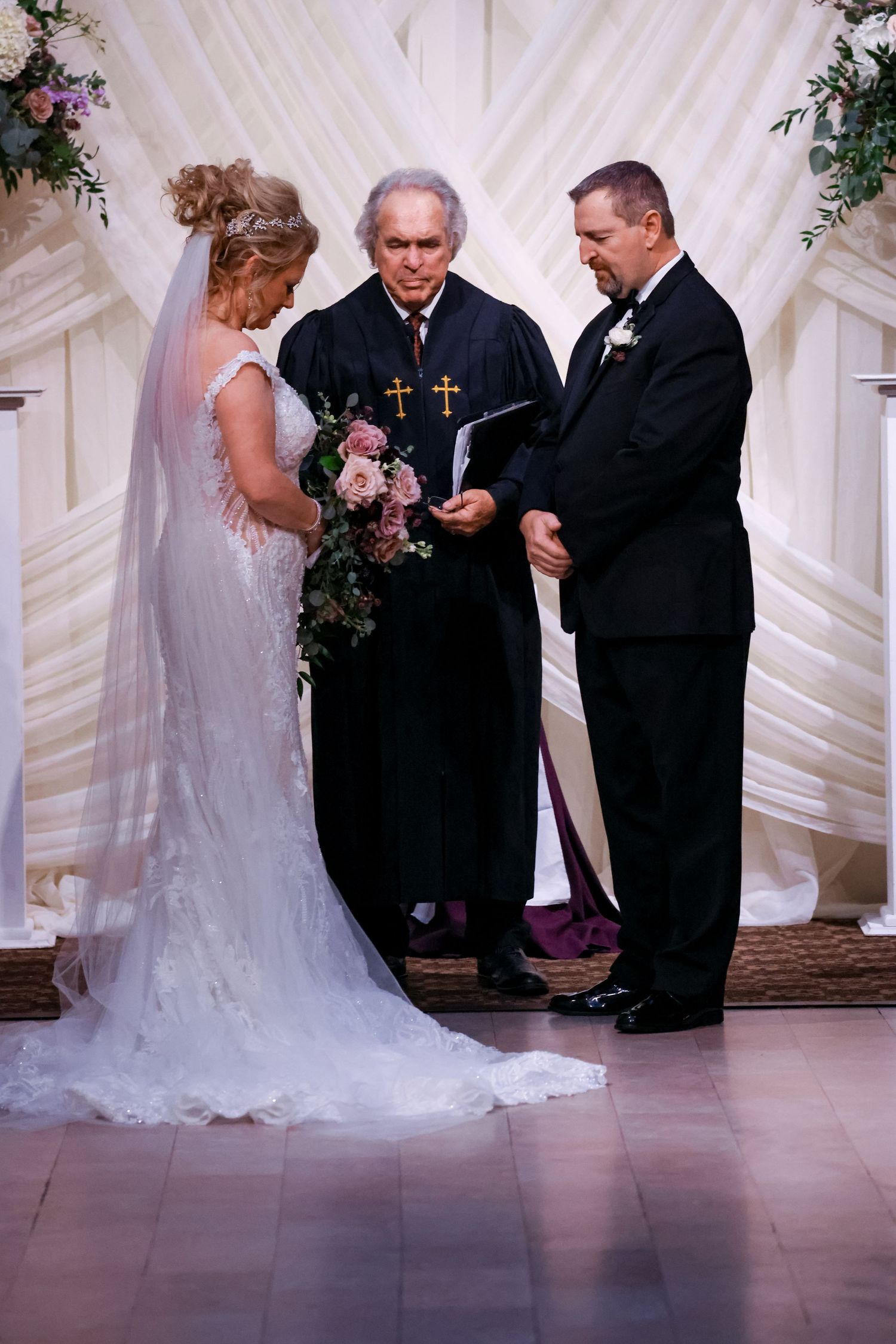 A traditional wedding ceremony takes place with a minister officiating as a couple exchanges vows under white decor.