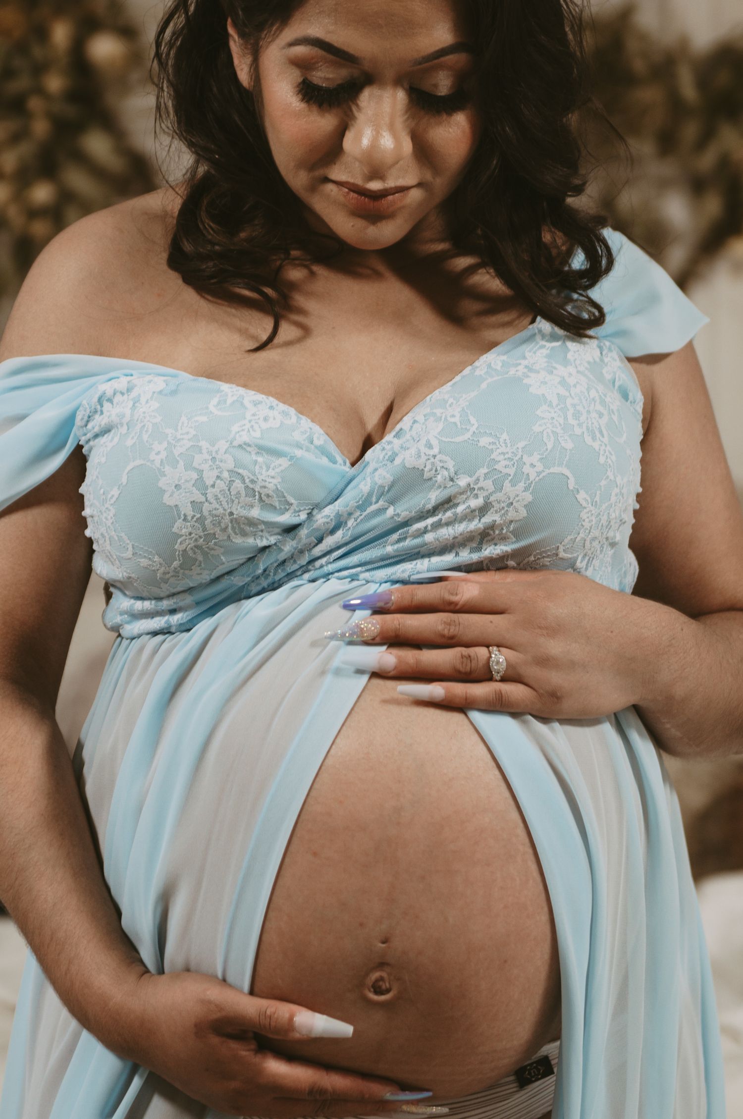 Maternity portrait in a light blue lace dress showing a baby bump against a natural backdrop.