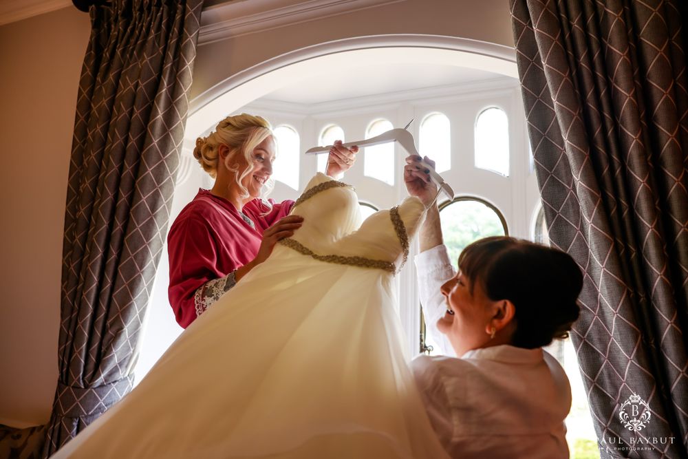 Wedding dress being prepared with care in an elegant window-lit room with dramatic curtains.