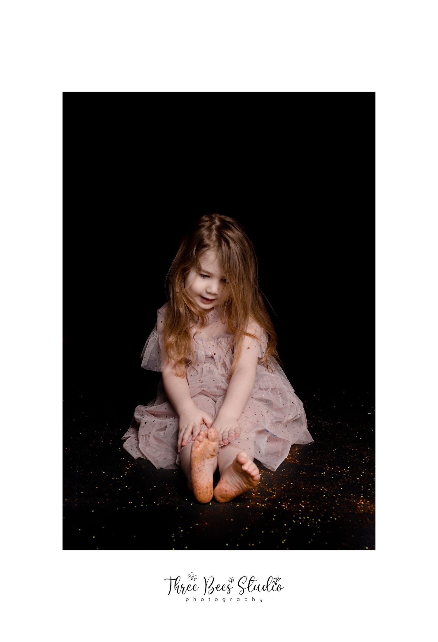Child in pink dress sitting on dark floor with glitter scattered around.