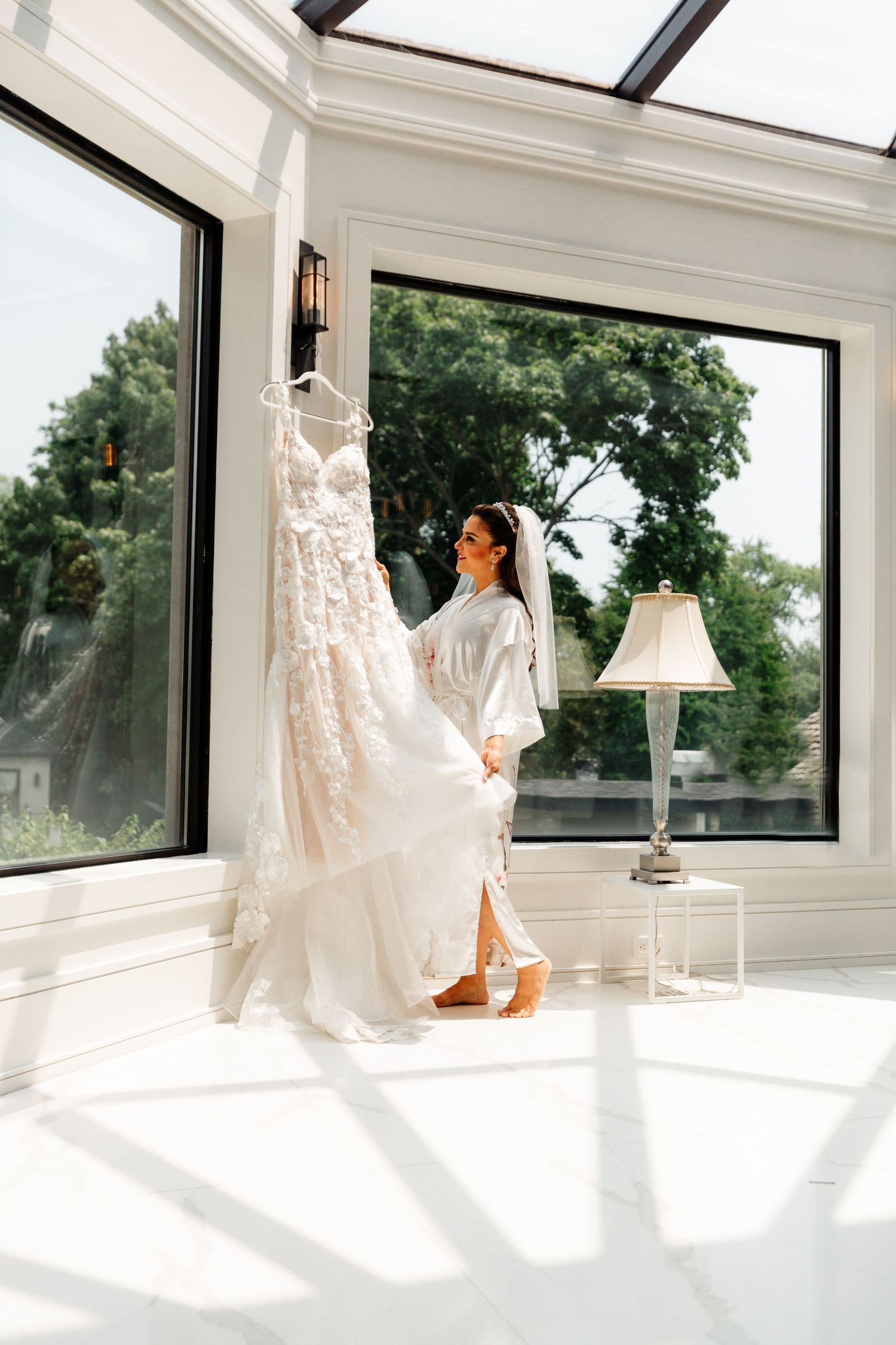 A wedding dress hangs elegantly in a sunlit room with large windows and a table lamp.