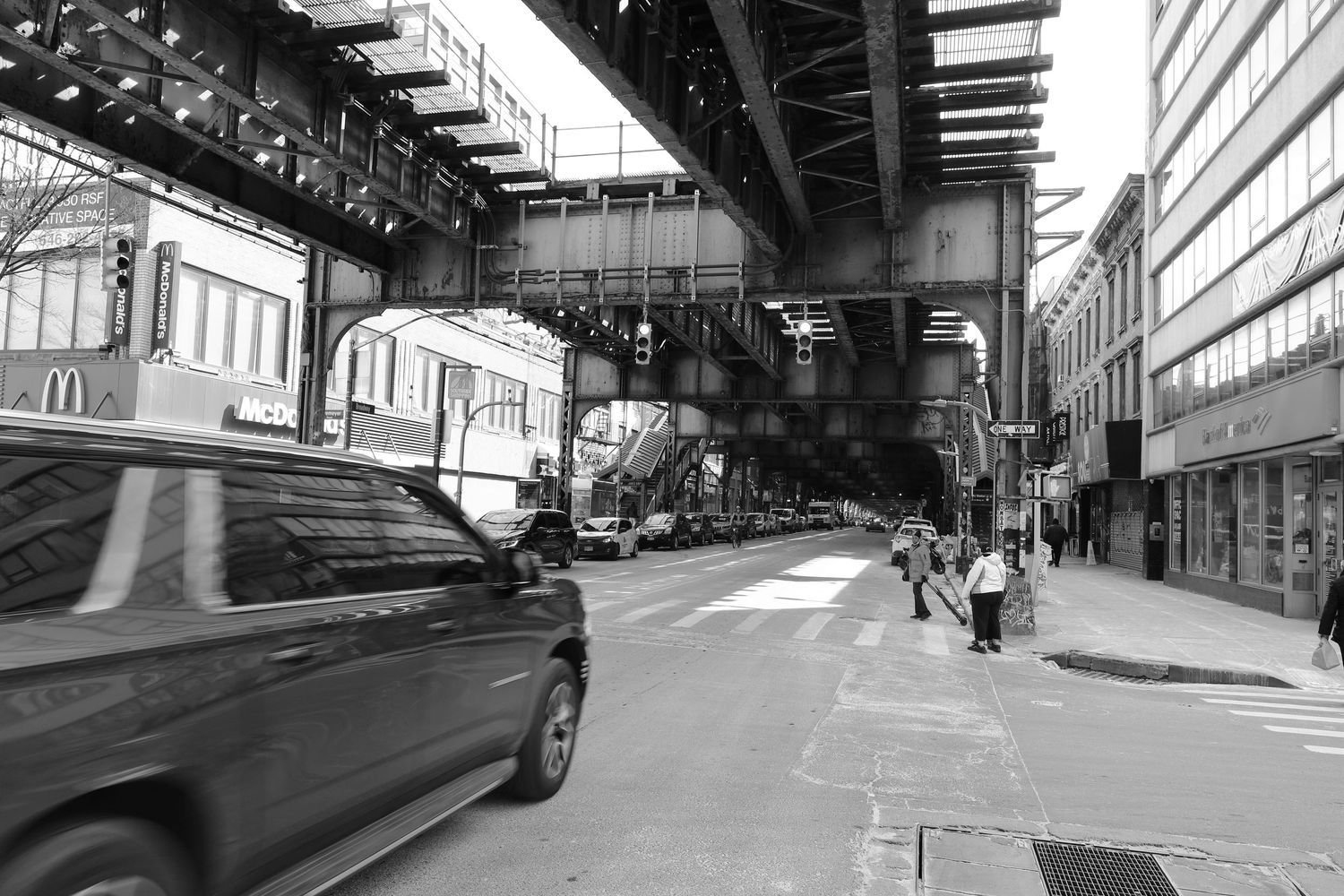 Black and white urban street scene under elevated train tracks with cars and pedestrians.