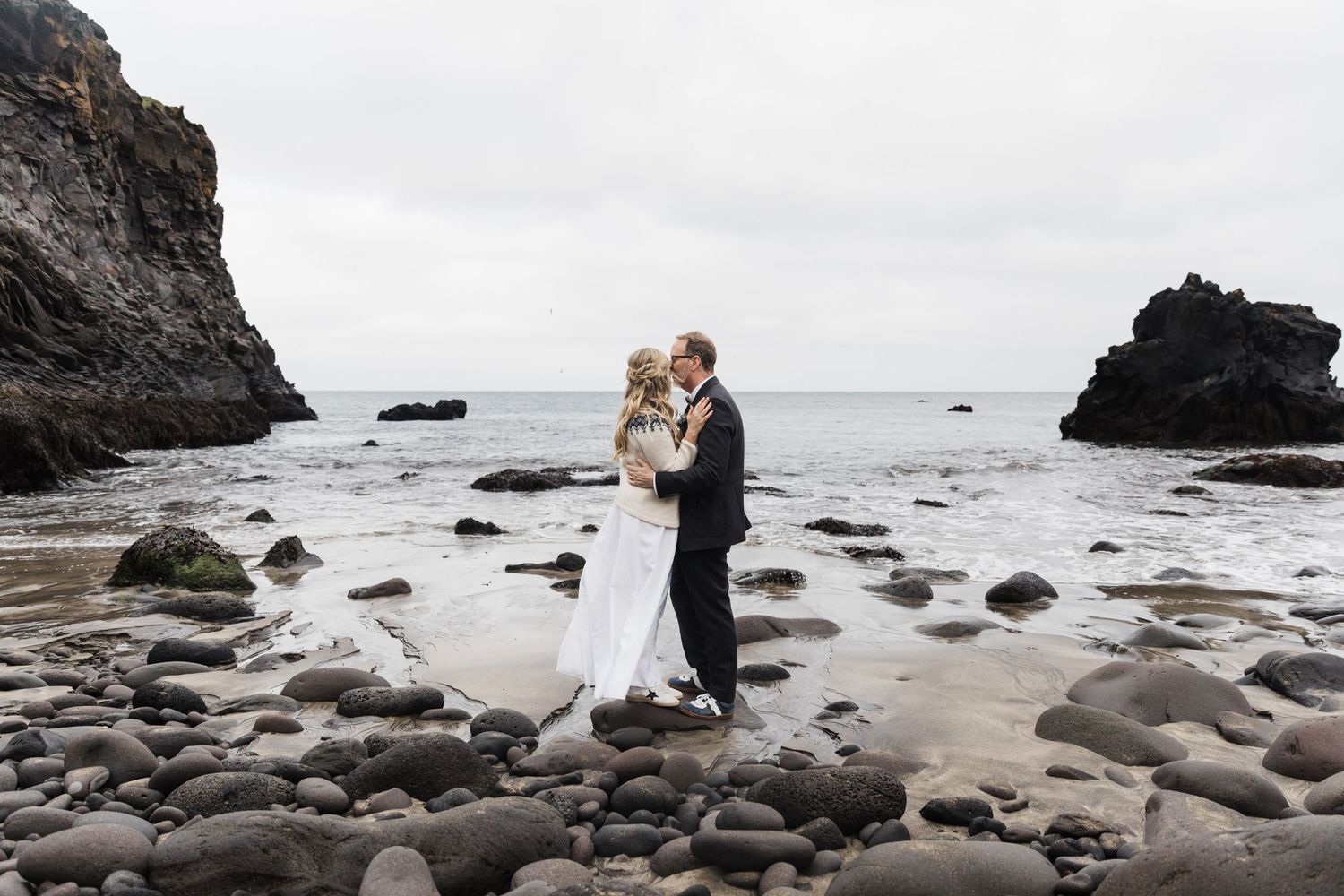 A romantic wedding moment on a rocky beach with dramatic cliffs and ocean waves in the background.