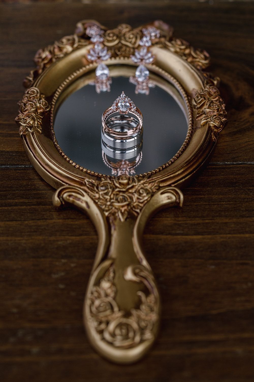 Gold ornate hand mirror with wedding bands, diamond engagement ring, and crystal drop earrings on wooden surface.