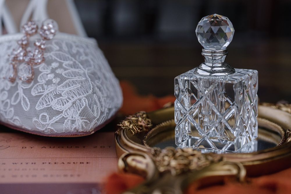 Crystal perfume bottle on gold tray beside lace bridal shoe and pink earrings, elegant wedding accessories flat lay.