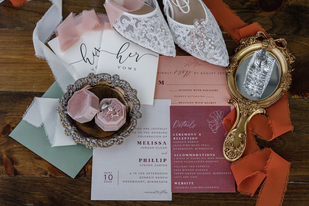 Elegant wedding flat lay featuring lace bridal shoes, pink velvet ring box, floral invitations, and a vintage gold mirror on wood.