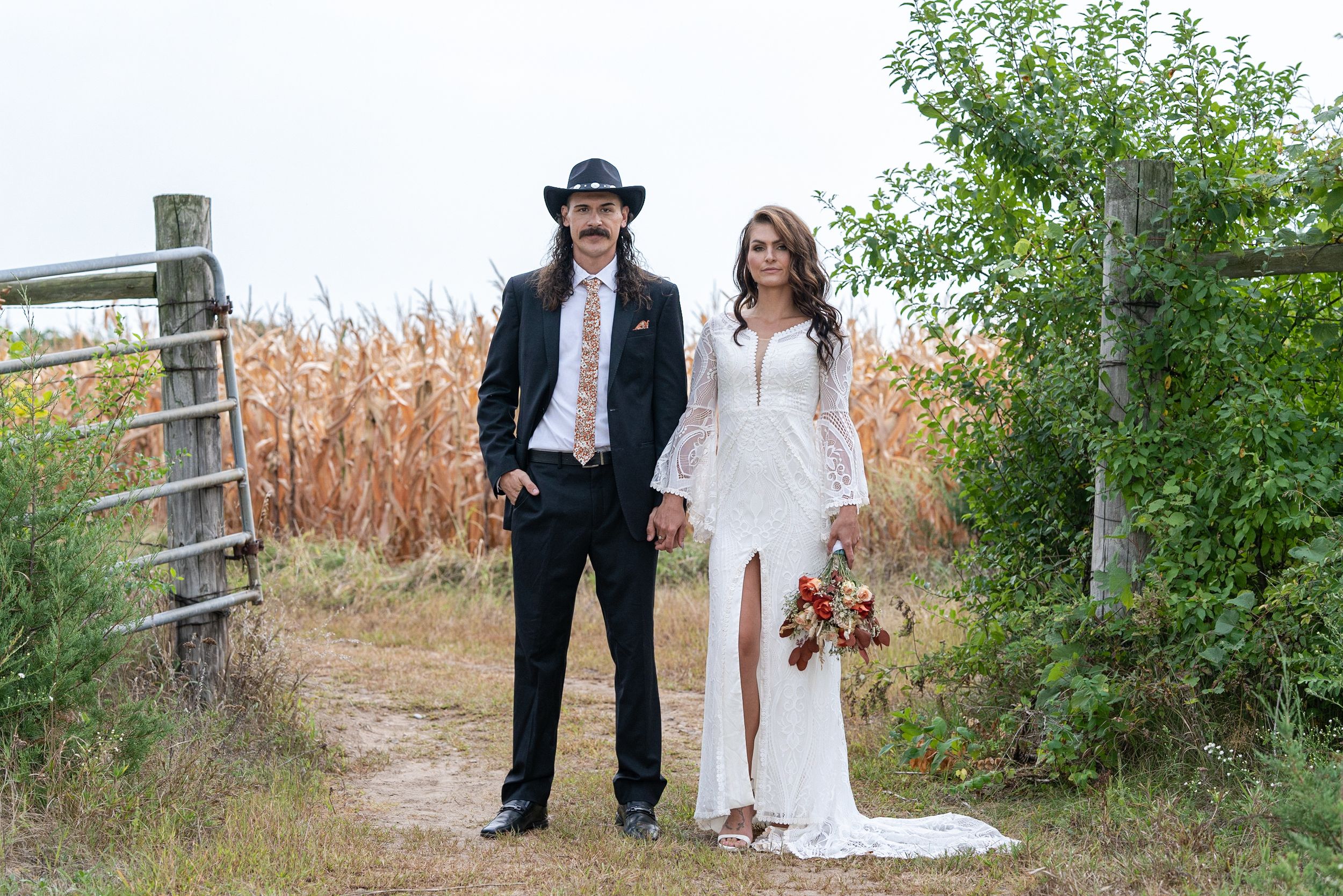 Bride in white lace gown and groom in black suit with cowboy hat pose on rustic dirt path near cornfield.