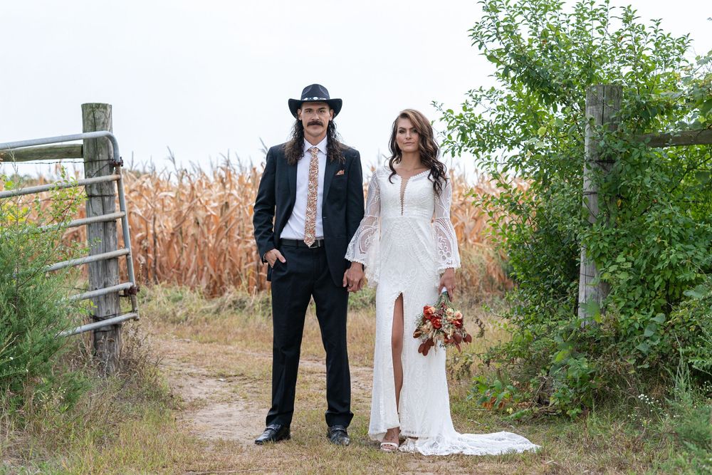 Bride in white lace gown and groom in black suit with cowboy hat pose on rustic dirt path near cornfield.