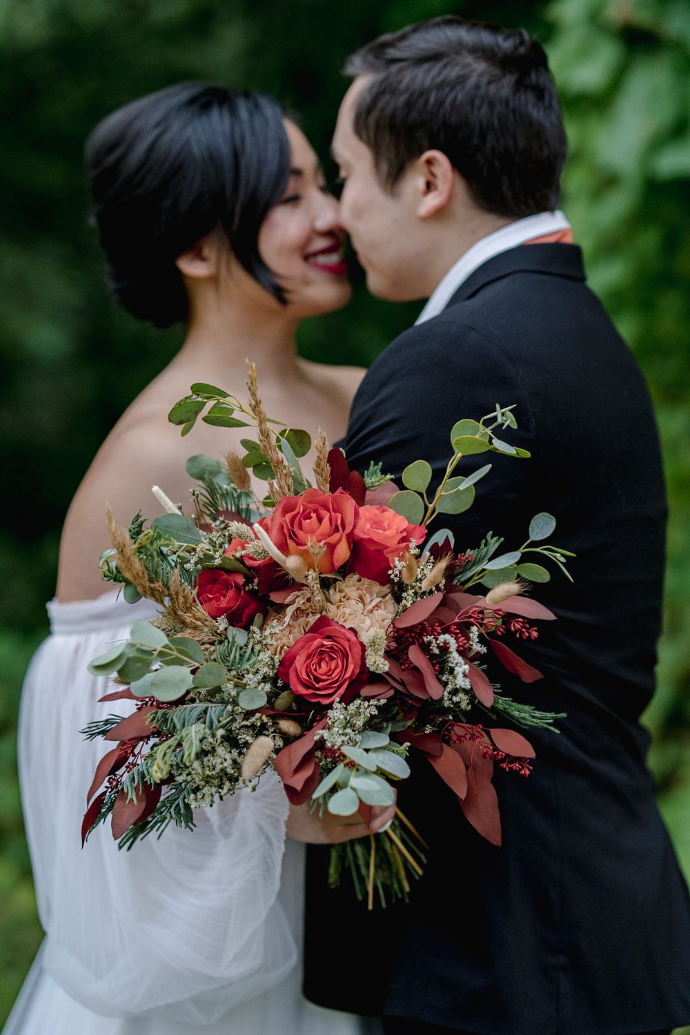Bride and groom sharing a romantic moment, holding a lush red rose and eucalyptus wedding bouquet outdoors.