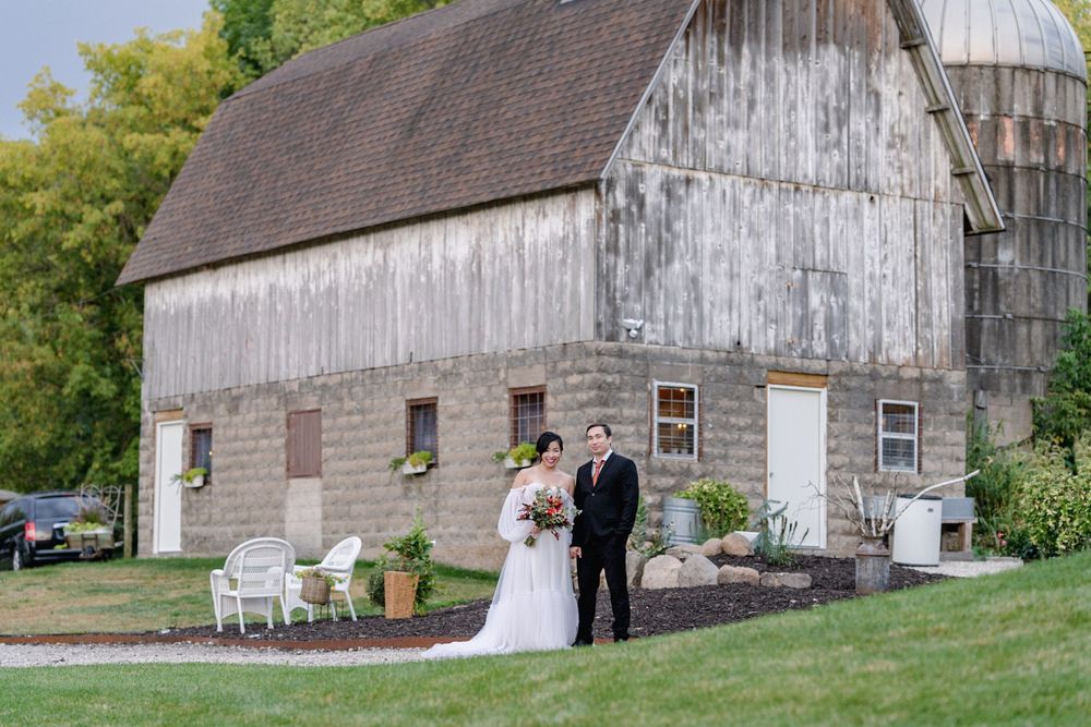 Bride and groom pose together in front of a rustic white barn on a lush green lawn at an outdoor wedding venue.