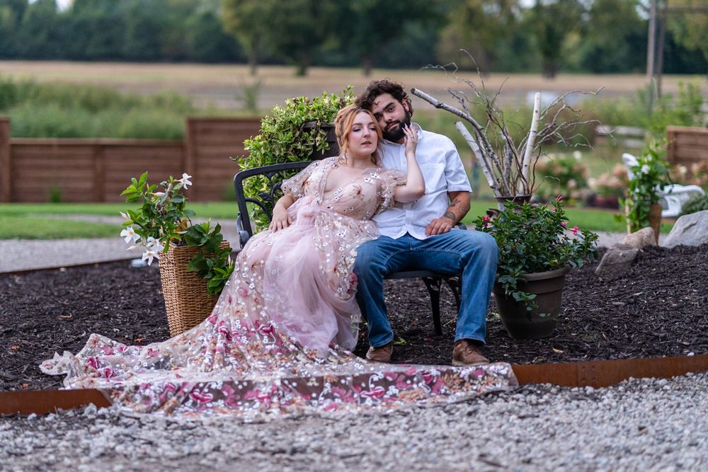 Couple posing in a garden, woman in floral gown resting on man in white shirt on a chair.