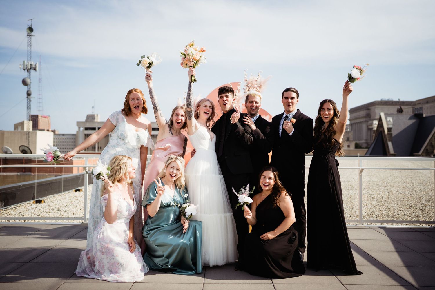 Wedding party celebrates on a rooftop with raised champagne glasses against an urban backdrop.